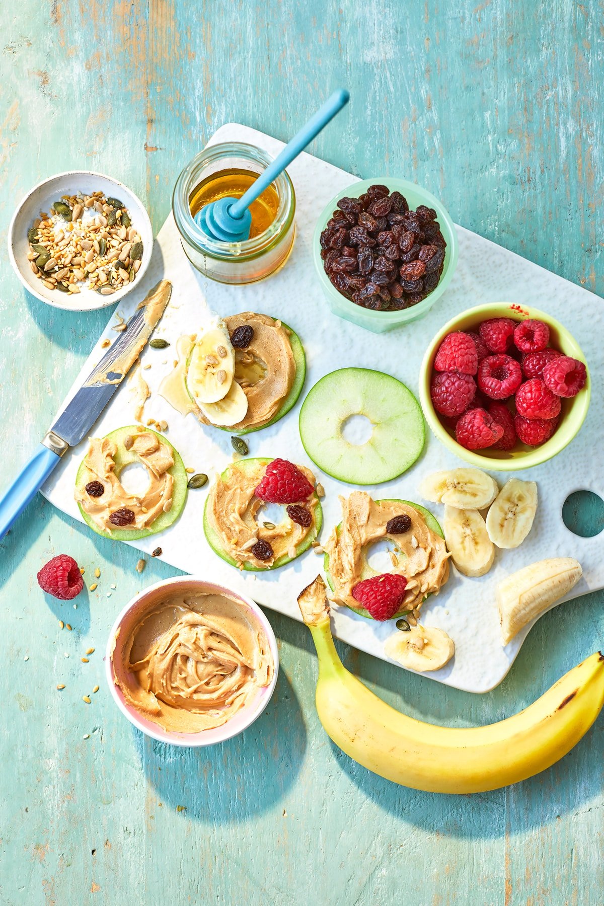 Apple slices with peanut butter on a white board, surrounded by topping ingredients such as raspberries, seeds, honey and banana slices.