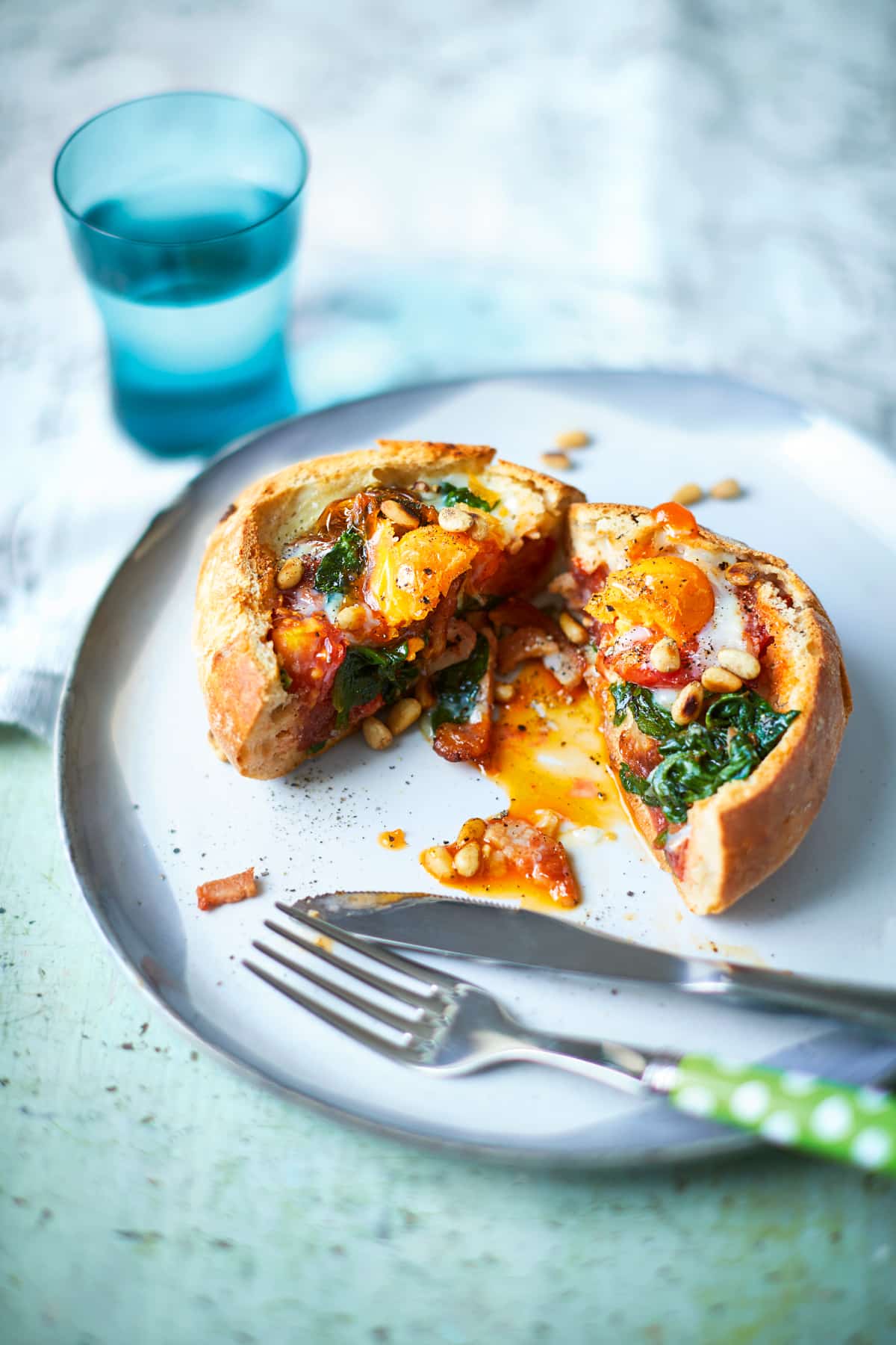 A breakfast roll, filled with sausage, bacon, spinach, tomatoes and pine nuts, topped with an egg. The roll is cut open to reveal the runny egg yolk. A knife and fork rest on the white plate.