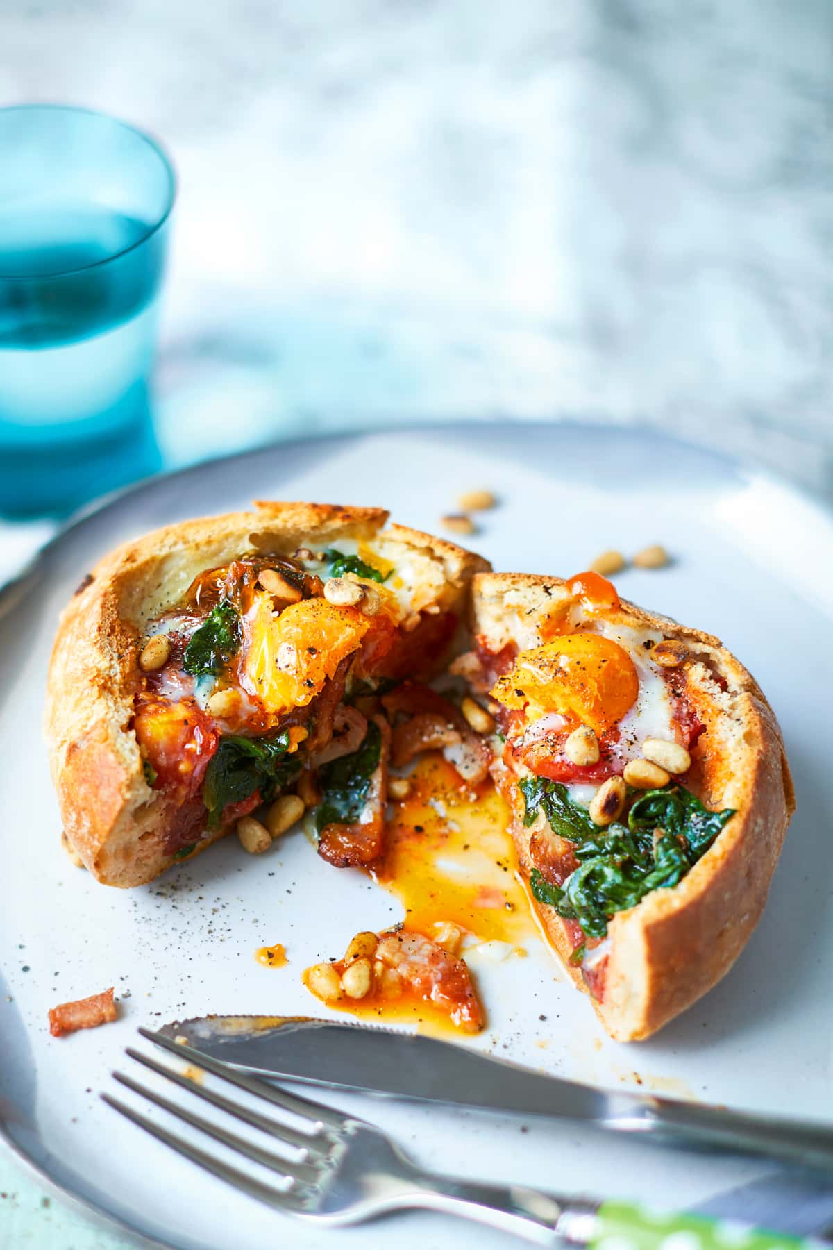 A cut open breakfast roll on a white plate. It is filled with sausage, bacon, spinach, tomatoes and pine nuts, topped with an egg. The roll is cut open to reveal the runny egg yolk. A glass of water is shown in the background.