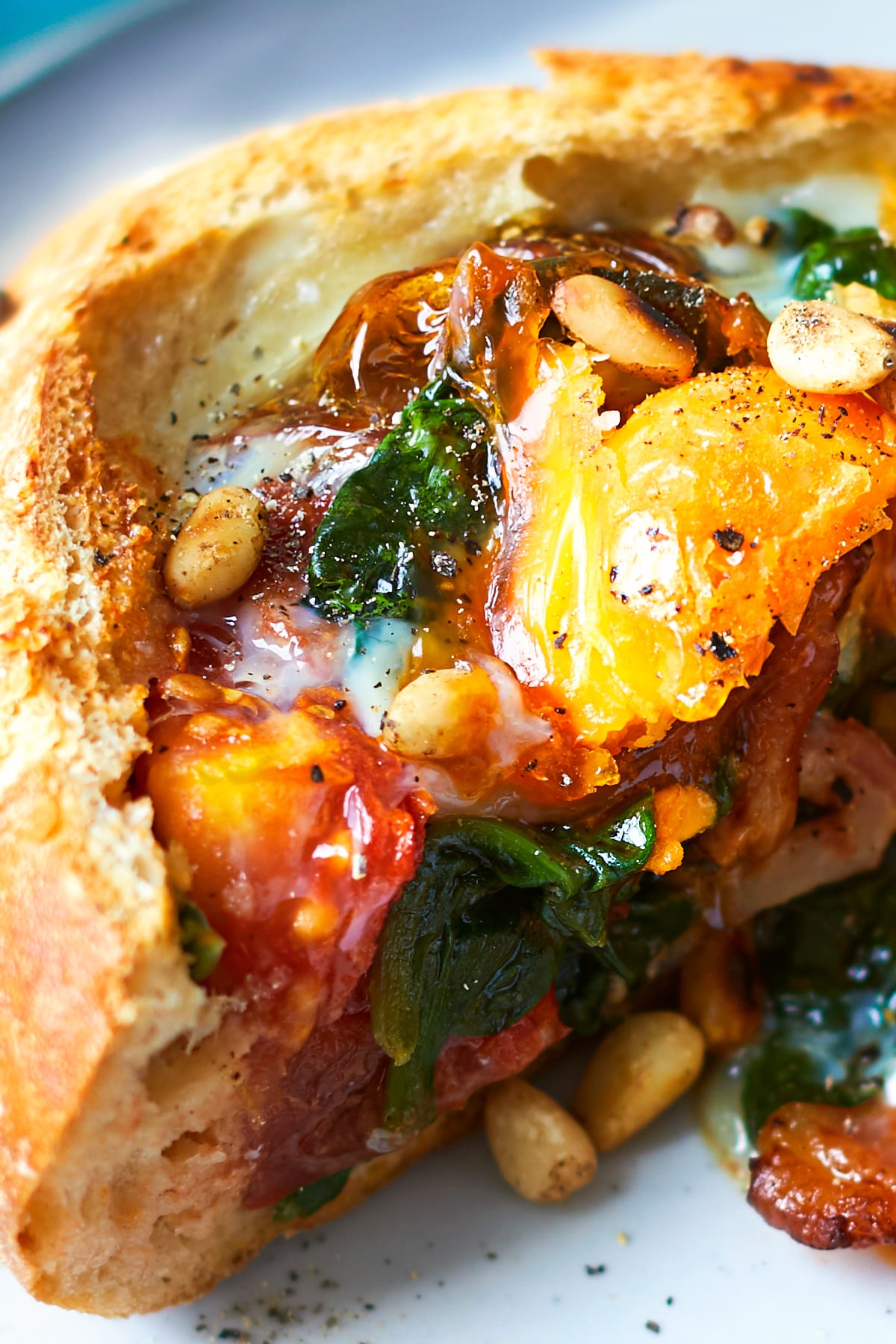 A cut open breakfast roll on a white plate. The close-up image shows that the roll is filled with sausage, bacon, tomatoes, spinach, tomatoes and pine nuts, topped with an egg. 