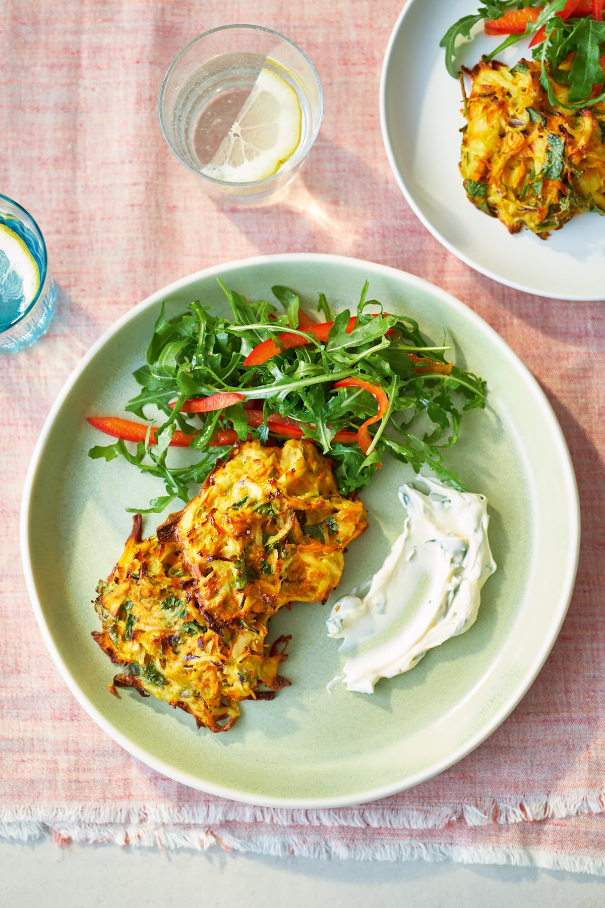 Two carrot patties on a green plate with a rocket and pepper salad. The plate sits on a pink background, with another plate shown, partially off-frame.