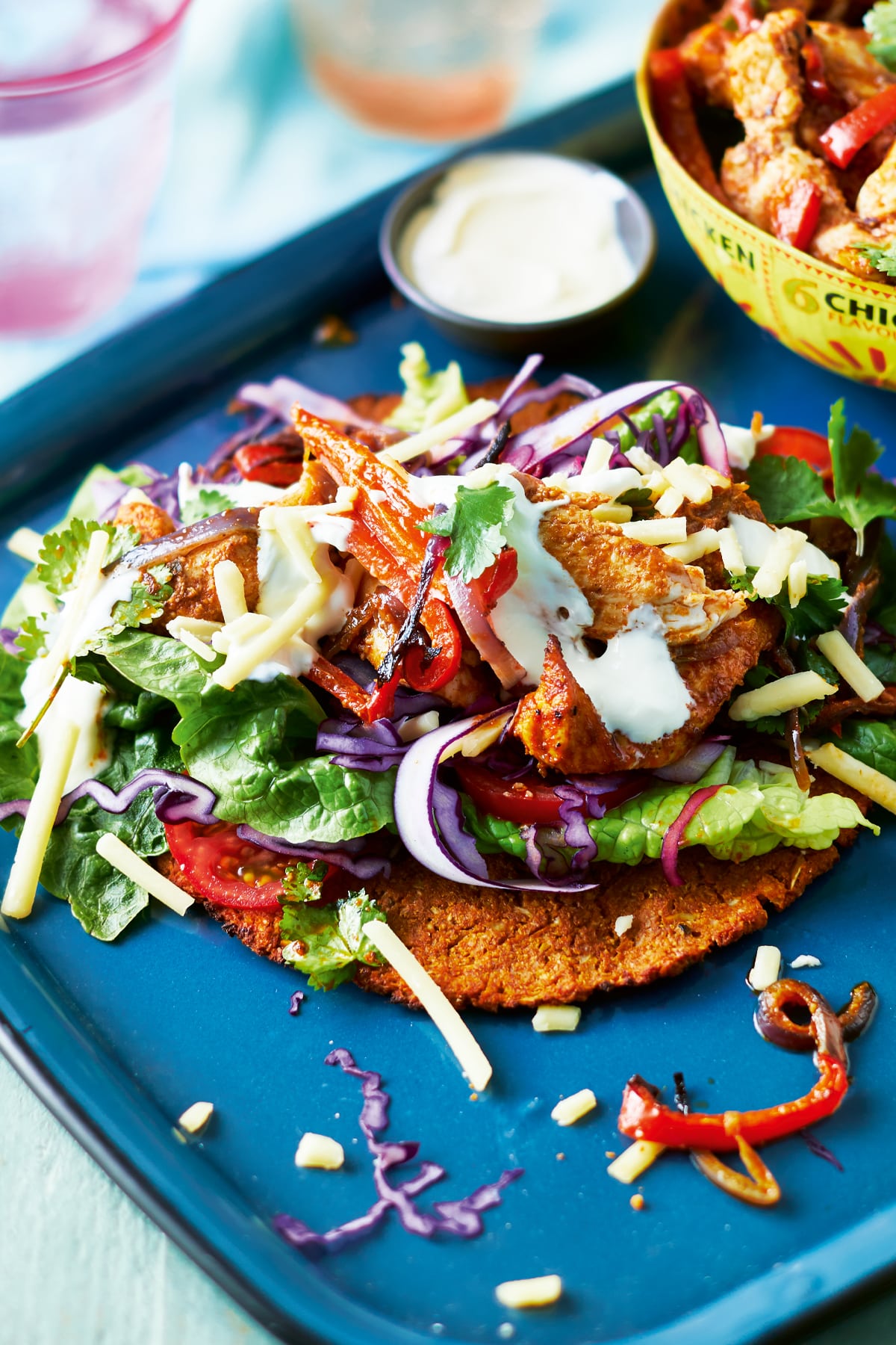 A cauliflower fajita with salad and chicken on a blue tray. Chicken and vegetable filling is shown in a yellow bowl in the background. 