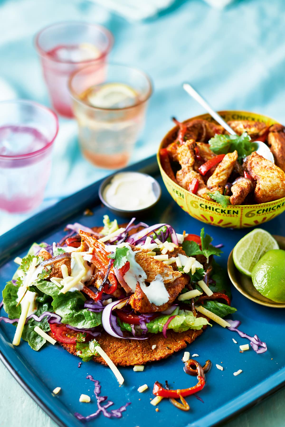 A cauliflower fajita packed with chicken and salad on a blue tray. Chicken and vegetable filling is shown in a yellow bowl, alongside cut limes. 