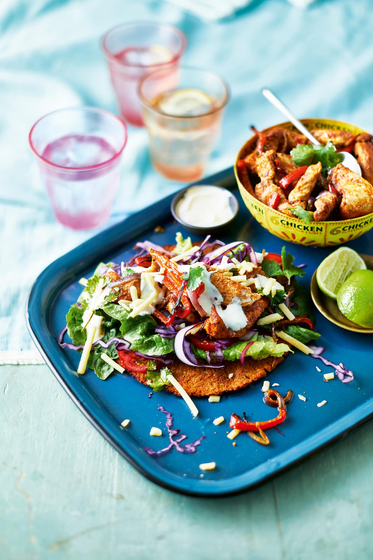 A cauliflower fajita with chicken and salad on a blue tray. Chicken and vegetable filling is shown in a yellow bowl, alongside cut limes. Glasses of water sit in the background.