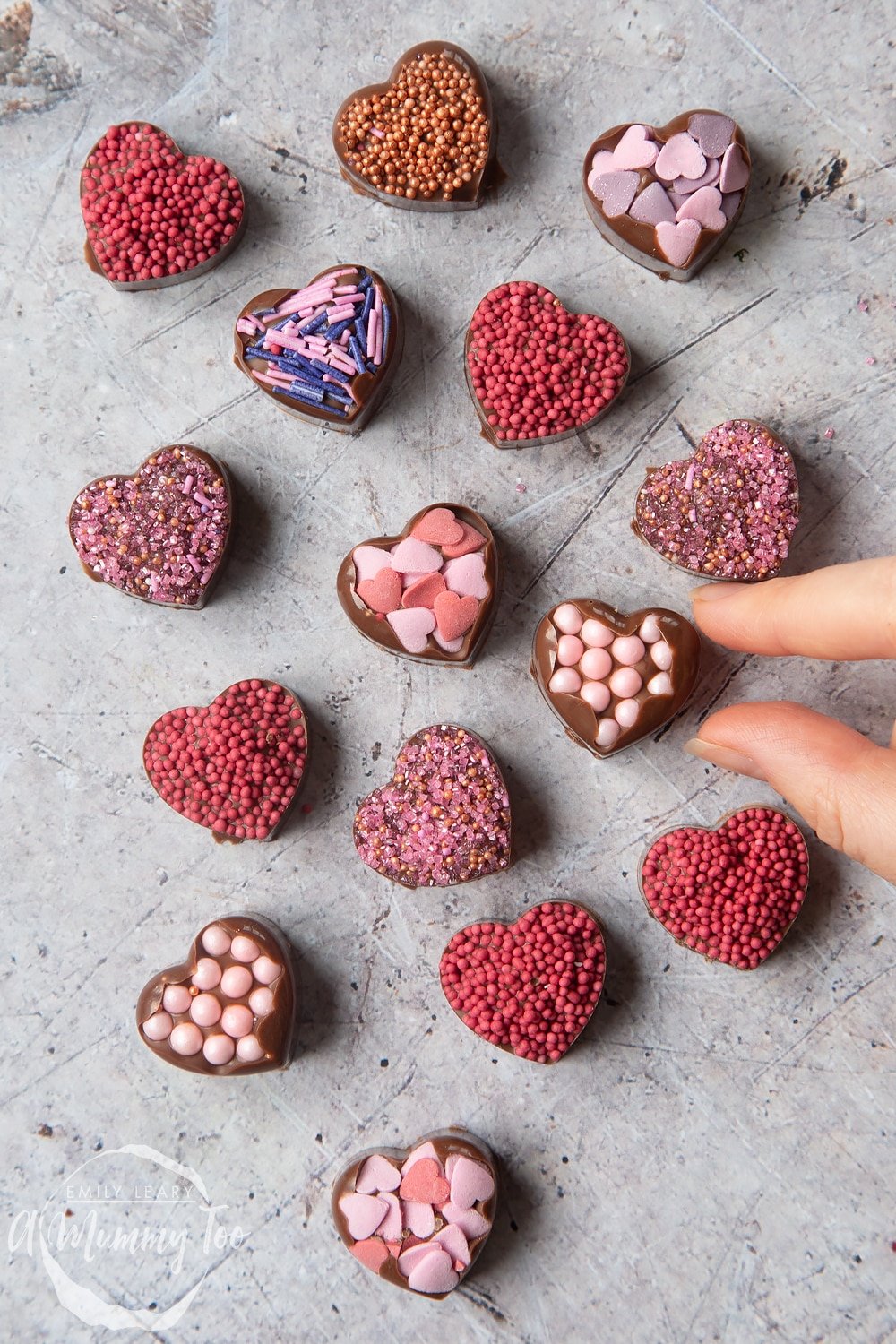 Overhead shot of a hand touching a chocolate heart with pink sprinkles with a mummy too logo in the lower-left corner