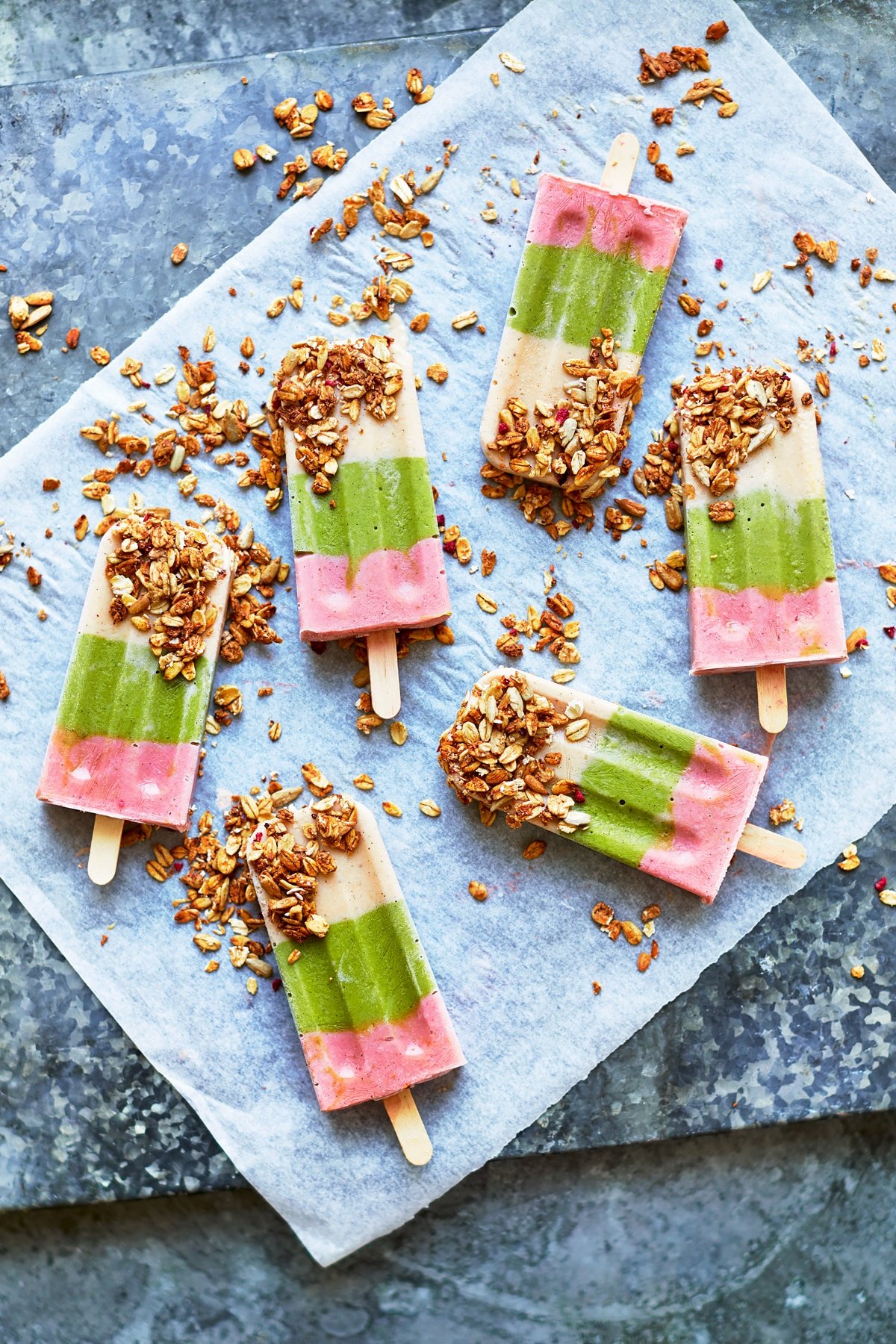 Fruit and veg lollies arranged on a piece of baking paper on a granite board. The lollies have been dipped in granola.