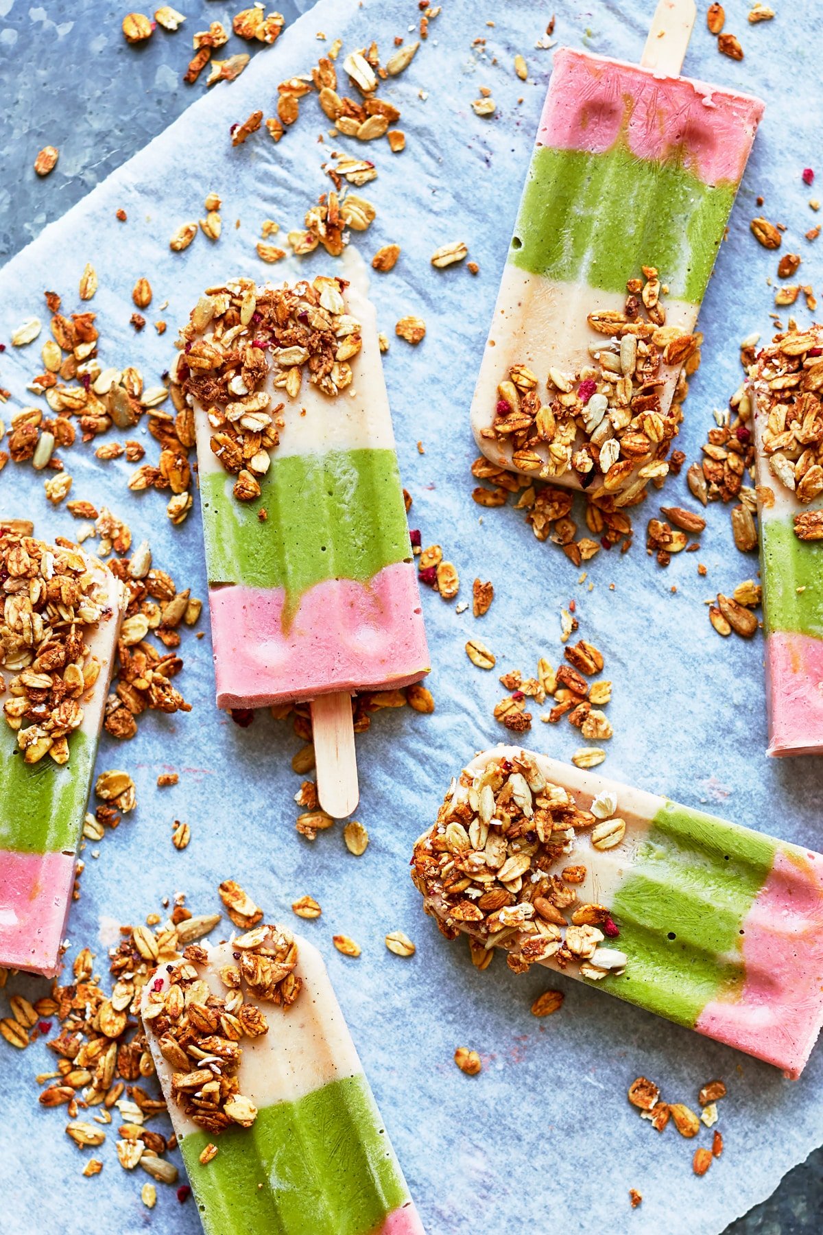 A close up of some fruit and veg lollies arranged on a piece of baking paper on a granite board. The lollies have been dipped in granola.