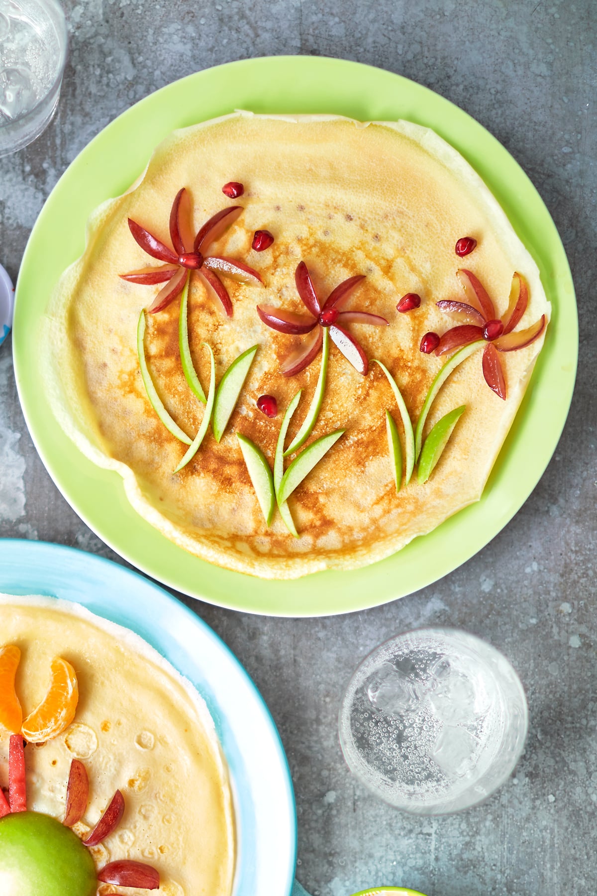 A crepe-style pancake on a green plate is decorated with fruit to resemble flowers.