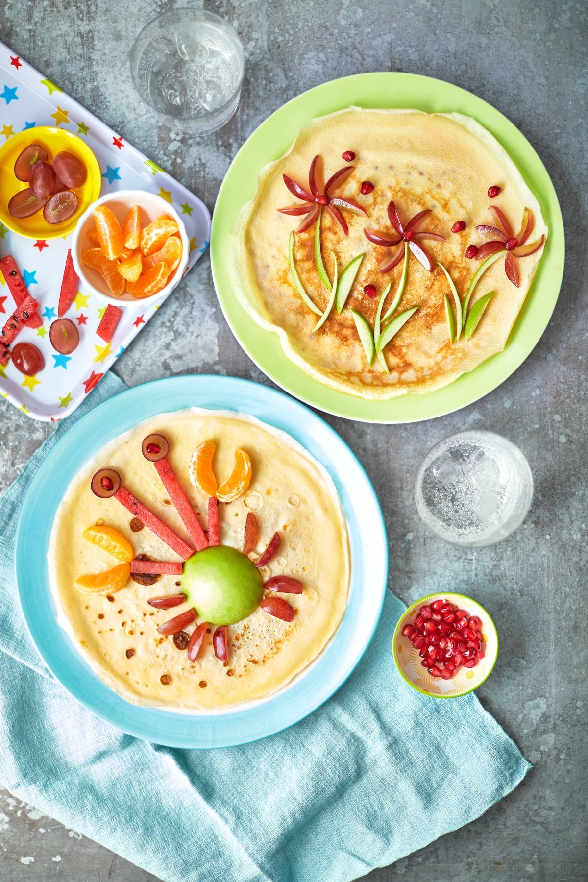 Two crepe-style pancakes on coloured plates. Both pancakes are decorated with fruit, one to resemble a crab and the other to resemble flowers.