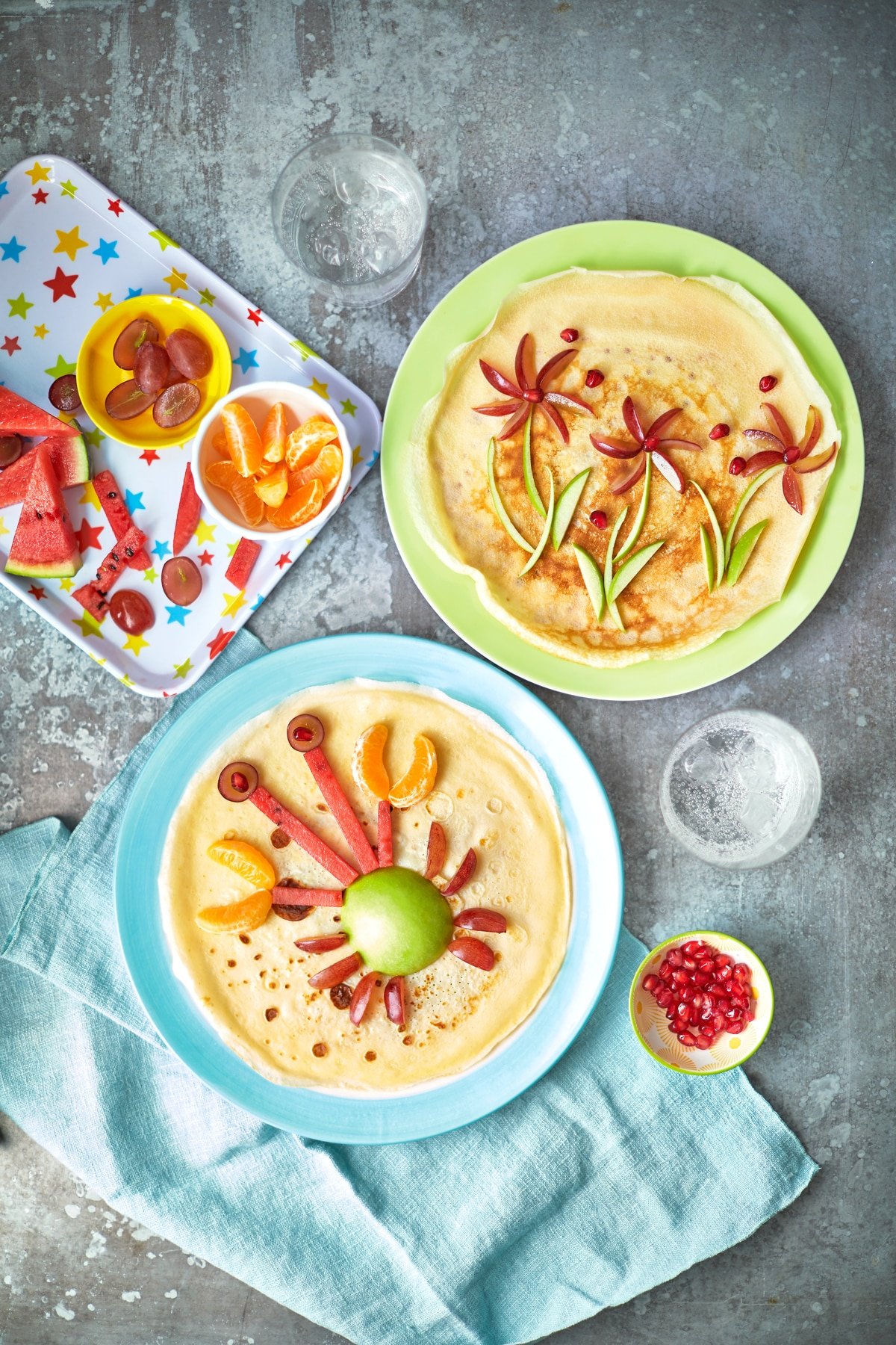 Two crepe-style pancakes on coloured plates. Both pancakes are decorated with fruit, one to resemble a crab and the other to resemble flowers. Further chopped fruit is shown on tray, to be used to create fun pancake ideas