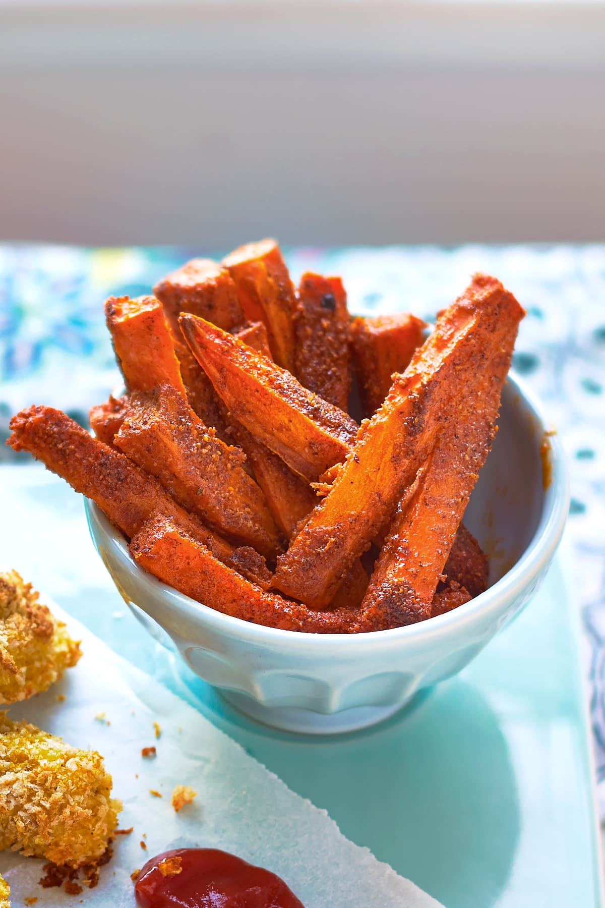 Sweet potato chips in a white bowl on a pale blue tray. Fish fingers are shown to the side.