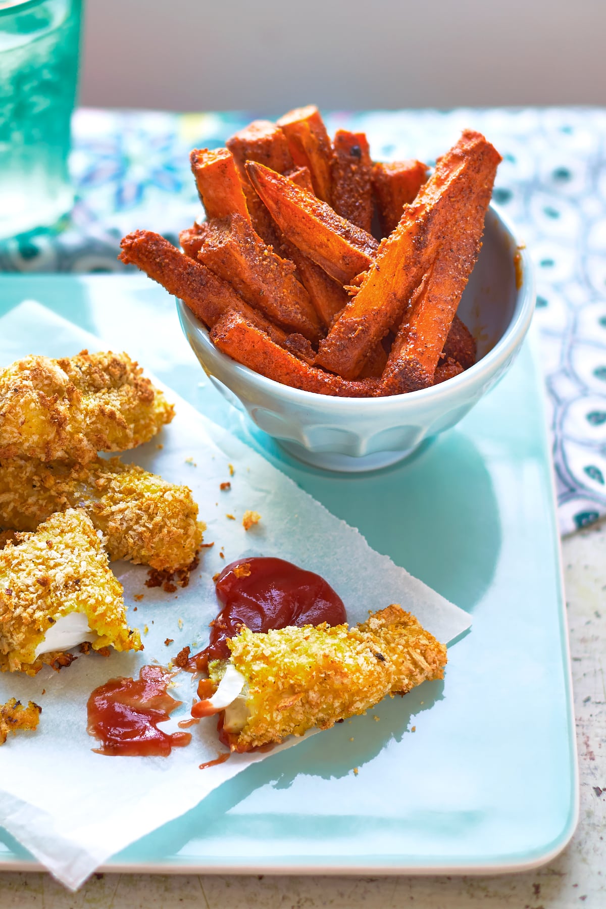 Sweet potato chips on a white bowl on a pale blue tray beside ketchup and spicy fish sticks.