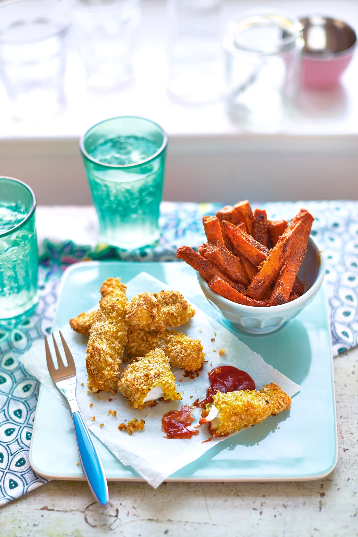 Sweet potato chips on a white bowl on a pale blue tray beside ketchup and fish sticks.