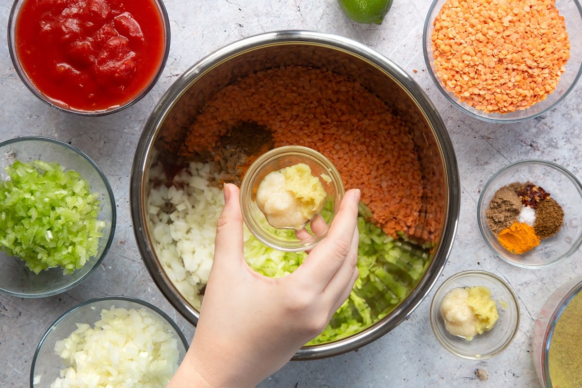 Overhead shot of a hand holding garlic and ginger in a small clear bowl above a slow cooker metal bowl 