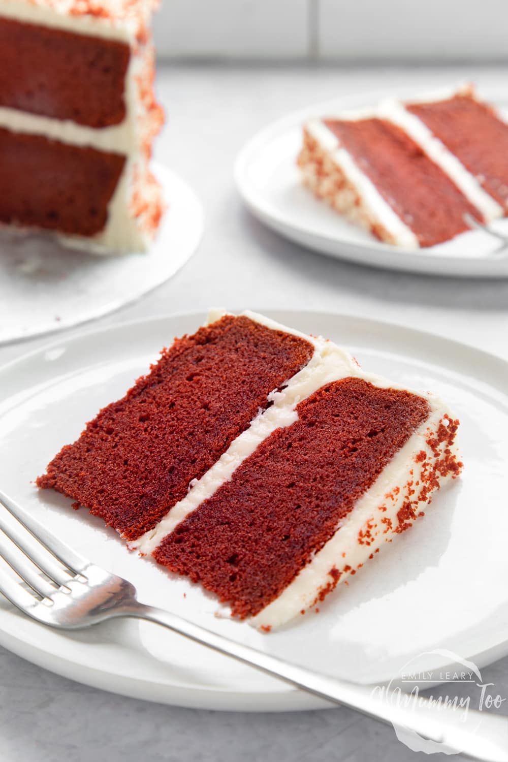 A slice of filled red velvet cake on a white plate. A fork rests on the plate. More cake is shown in the background.