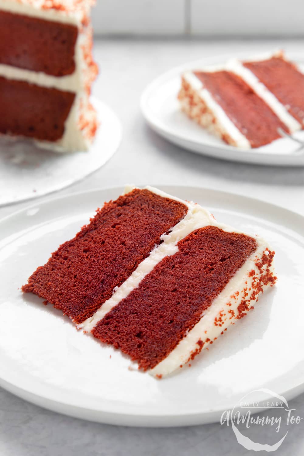 A slice of filled red velvet cake on a white plate. More cake is shown in the background.