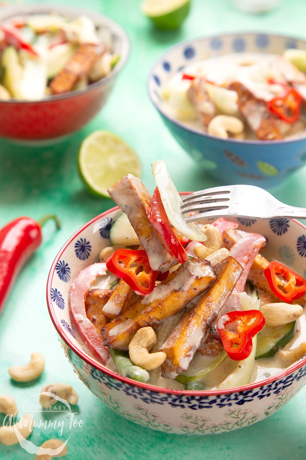 a fork picking up vegetables from a Thai green curry in a decorative bowl with two bowls in the background.