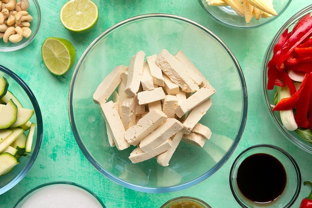 chopped slices of tofy in a clear mixing bowl surrounding by bowls of vegetables and ingredients.