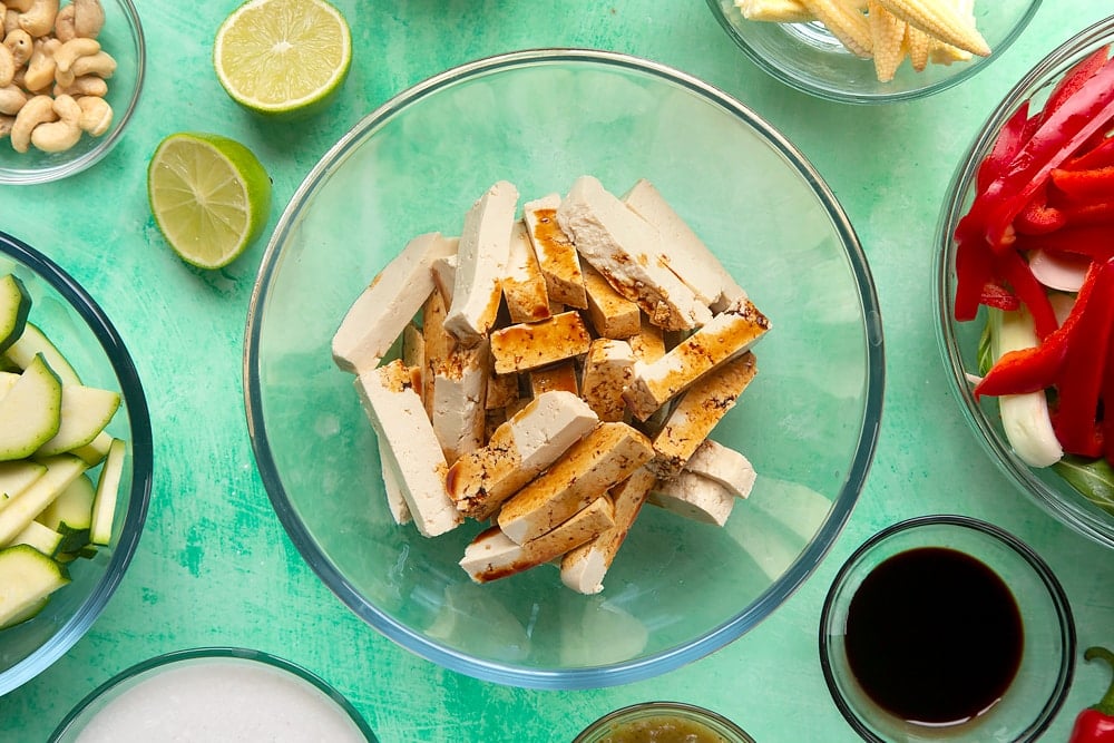 chopped strips of tofu covered in soy sauce in a clear mixing bowl surrounded by chopped vegetables