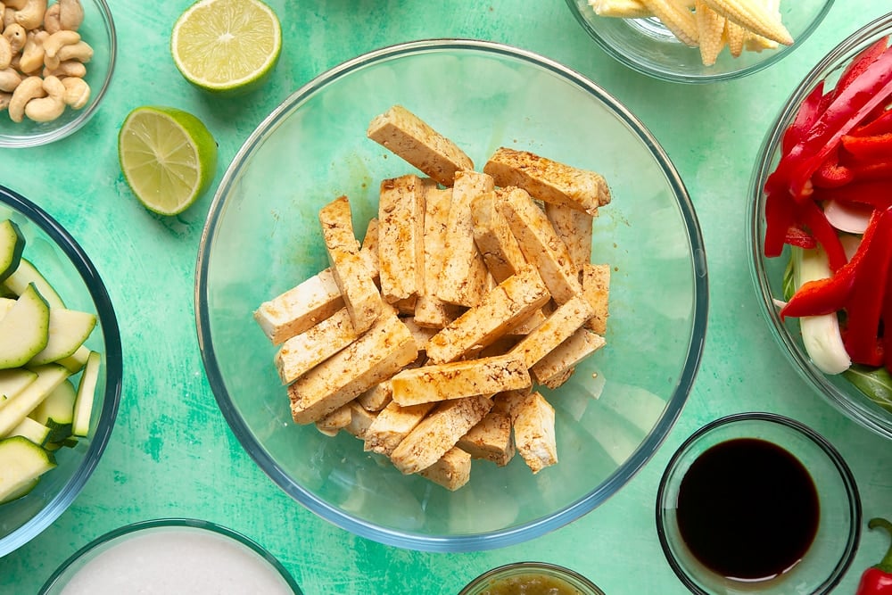 marinated tofu strips in a clear mixing bowl surrounded by sliced vegetables.