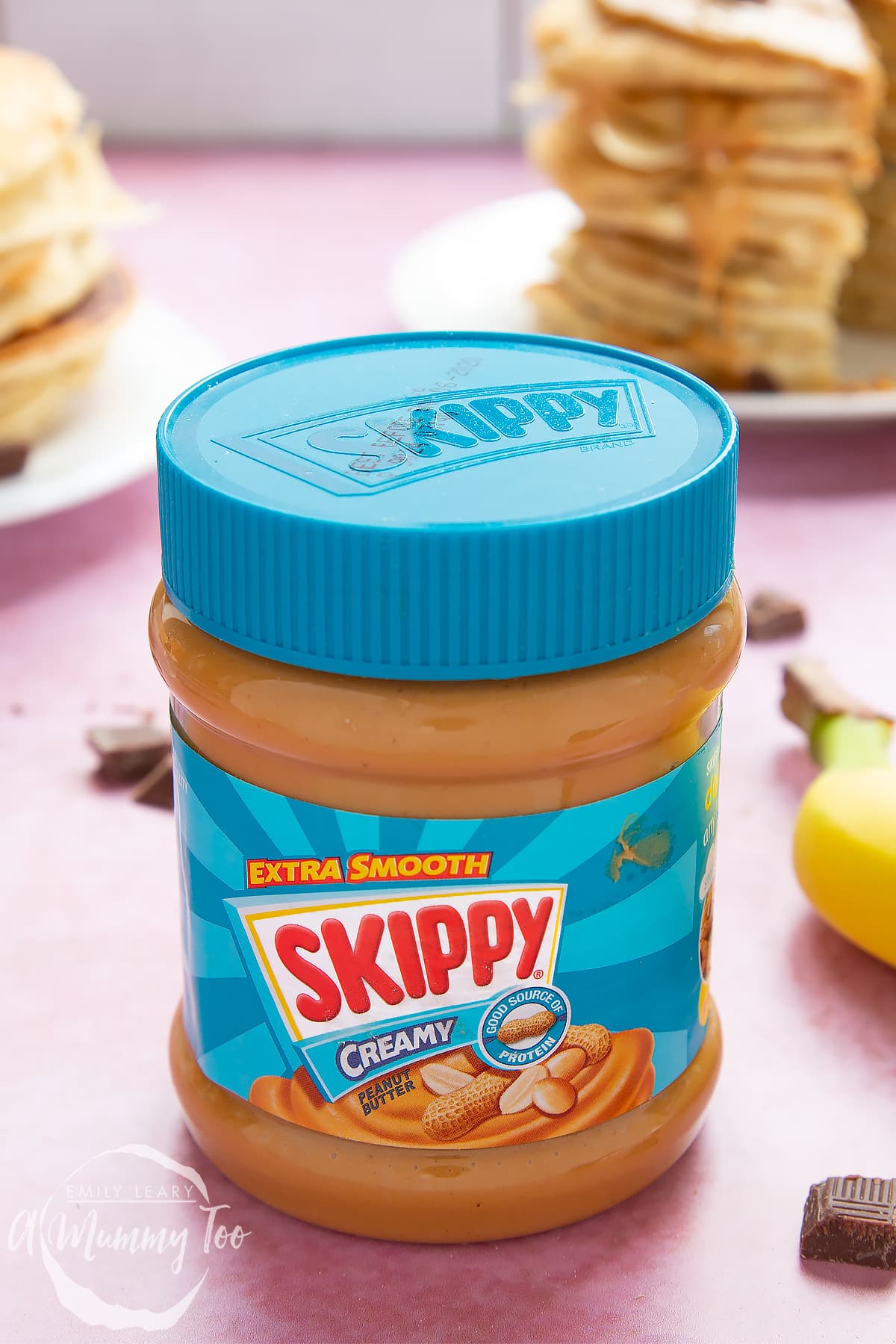 A jar of Skippy Smooth Peanut Butter. In the background are stacks of vegan peanut butter pancakes on white plates.