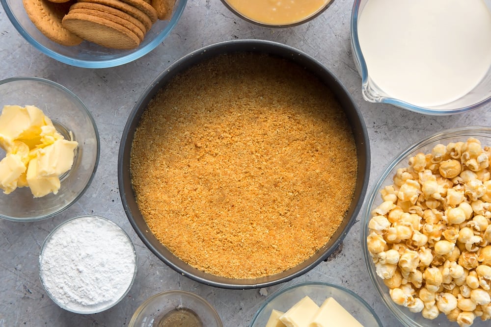 over head view of crushed biscuits and melted butter combined in cake tin surrounded by other ingredients.