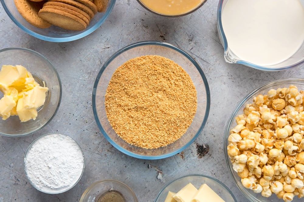 over head view of crushed digestive biscuits in a clear bowl in the centre of the table surrounded by ingredients in more clear bowls.