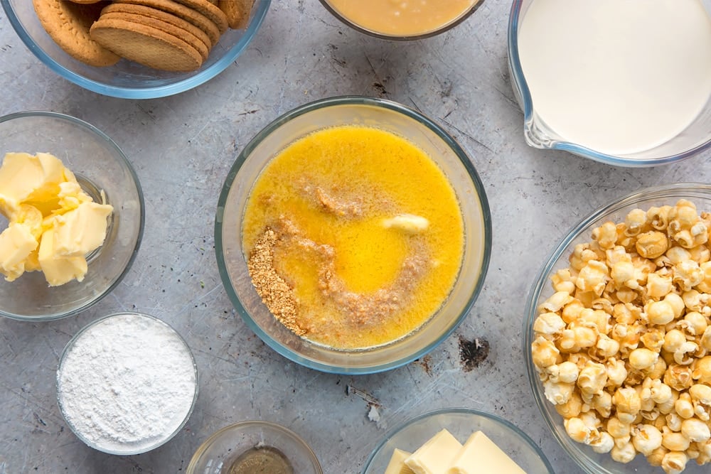 over head view of crushed biscuits and melted butter in a clear bowl surrounded by other ingredients.