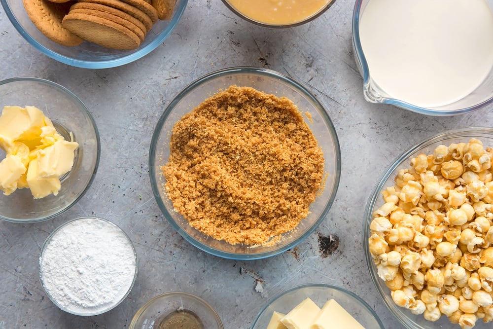 over head view of crushed biscuits and melted butter combined in a clear bowl surrounded by other ingredients.