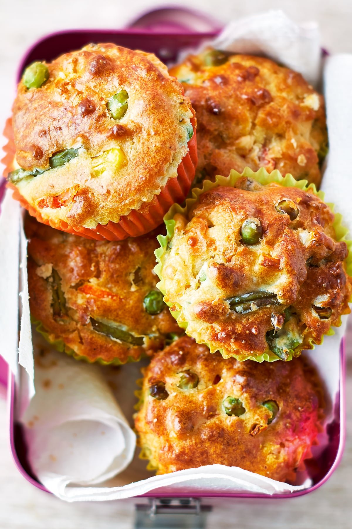 Savoury vegetable muffins in a pink lunchbox lined with paper. The muffins have golden brown tops, studded with peas and beans.