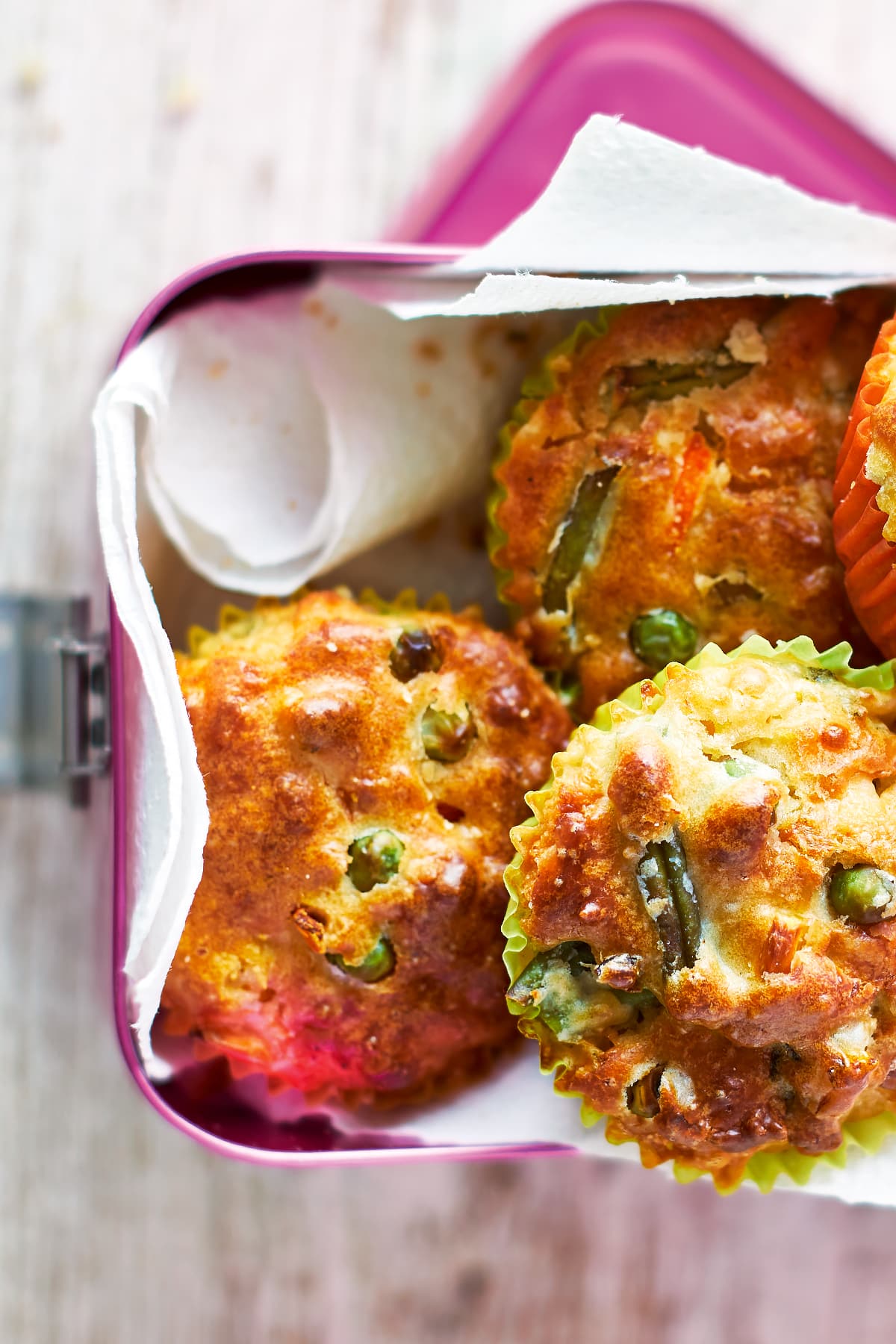 Savoury vegetable muffins in a pink lunchbox lined with paper. The muffins are in multicoloured cases and the tops are studded with carrots, peas, beans and sweetcorn.