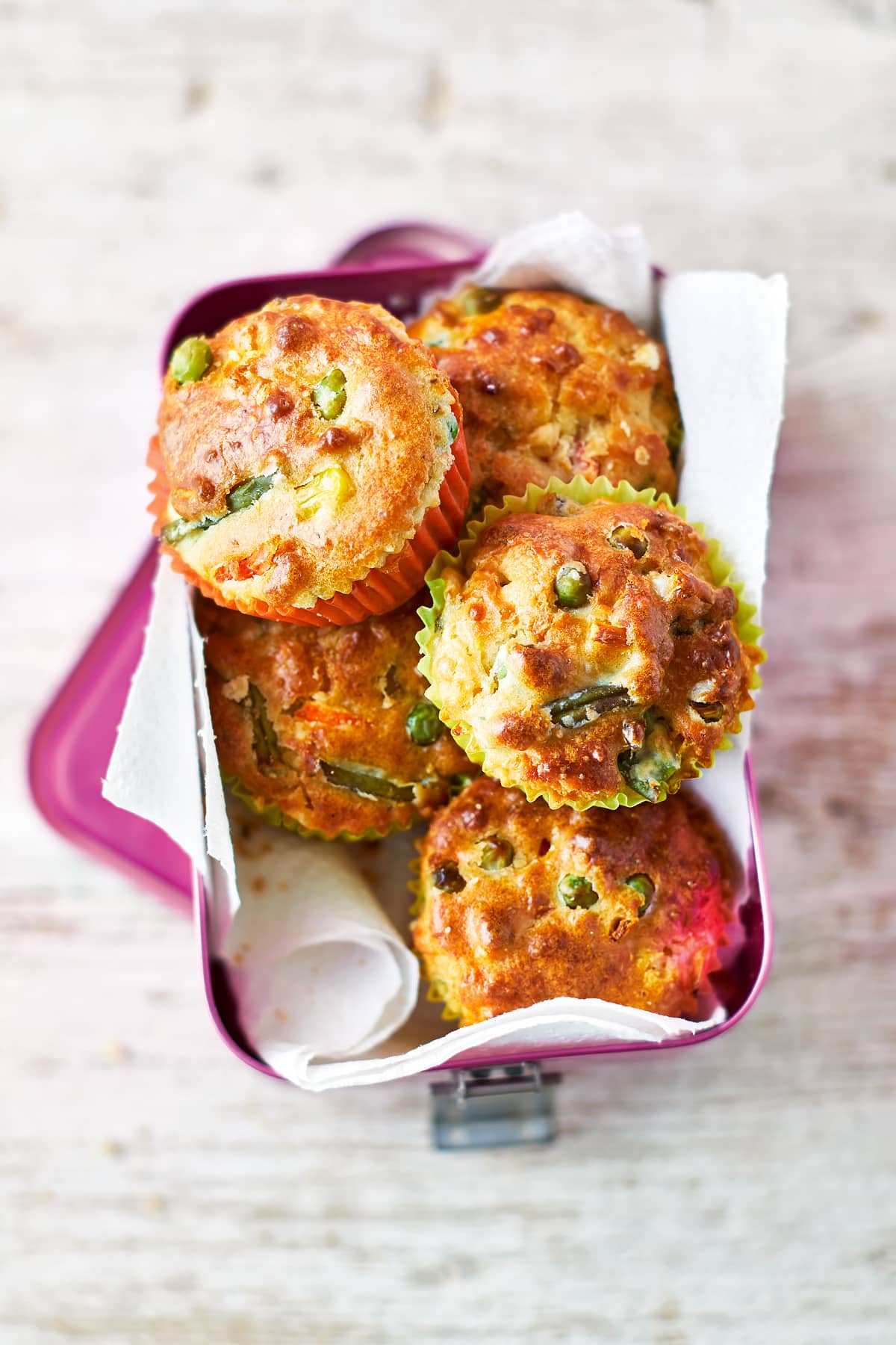 Savoury vegetable muffins in a pink lunchbox lined with paper on a wooden background. The muffins are in multicoloured cases and the tops are studded with carrots, peas, beans and sweetcorn.