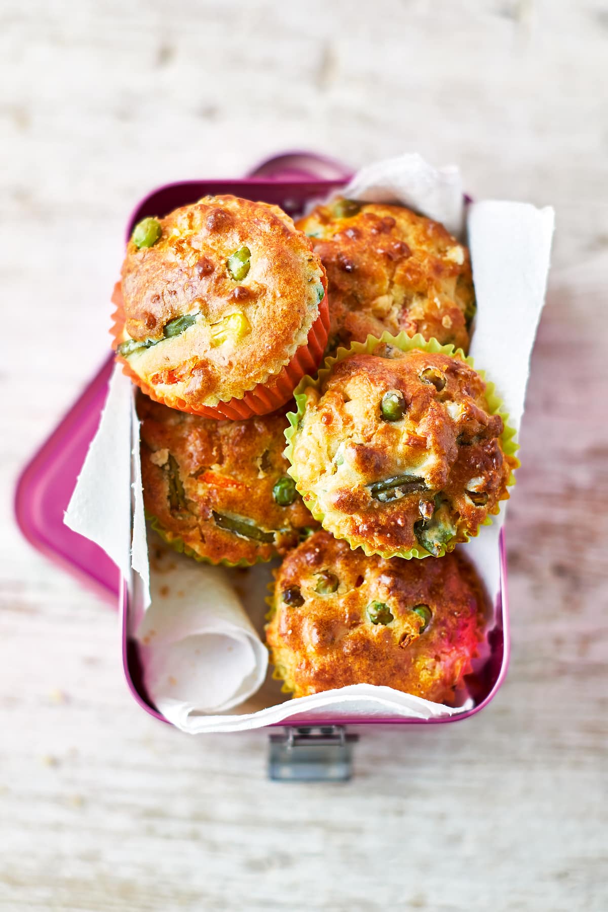 A pink lunchbox lined with paper, filled with savoury vegetable muffins. The muffins are in multicoloured cases and the tops are studded with carrots, peas, beans and sweetcorn.