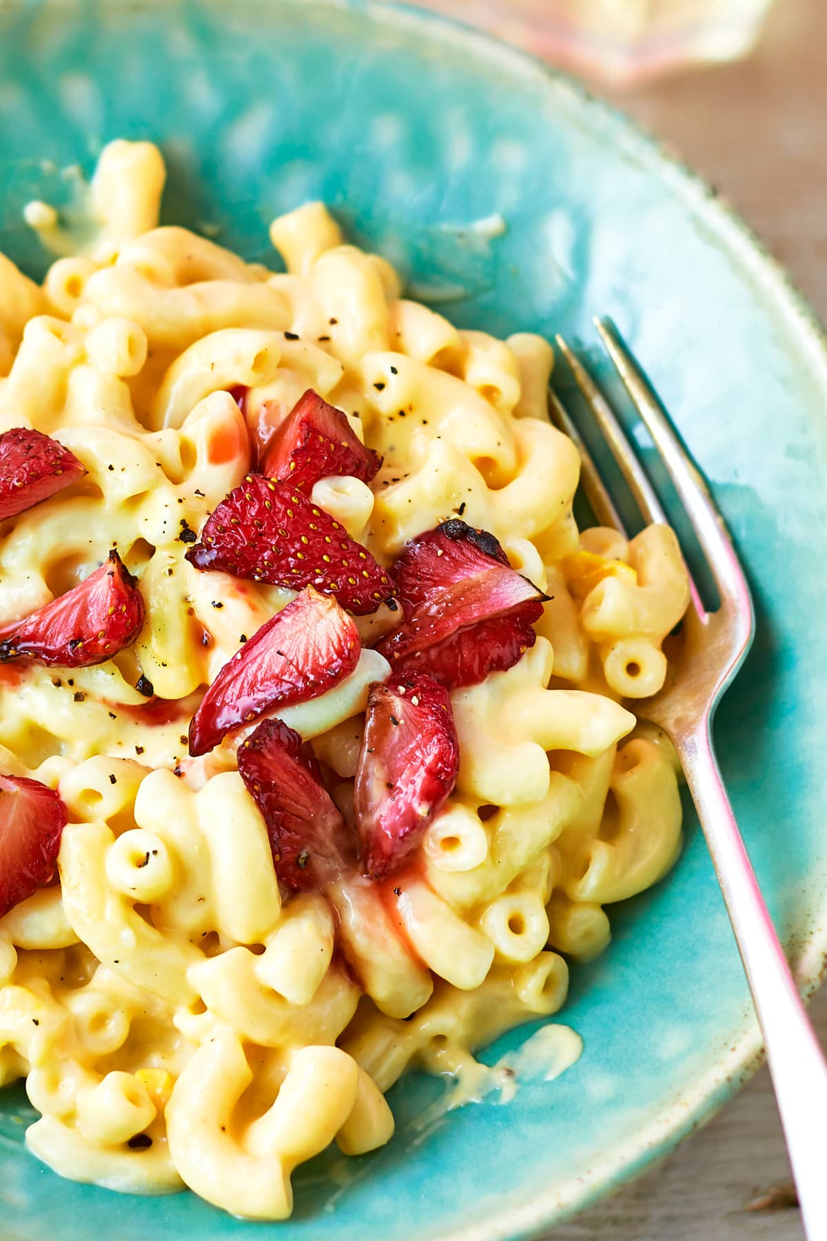 A turquoise bowl of strawberry pasta: macaroni in a cheese and butternut squash sauce, topped with roasted balsamic strawberries. A fork rests in the bowl.