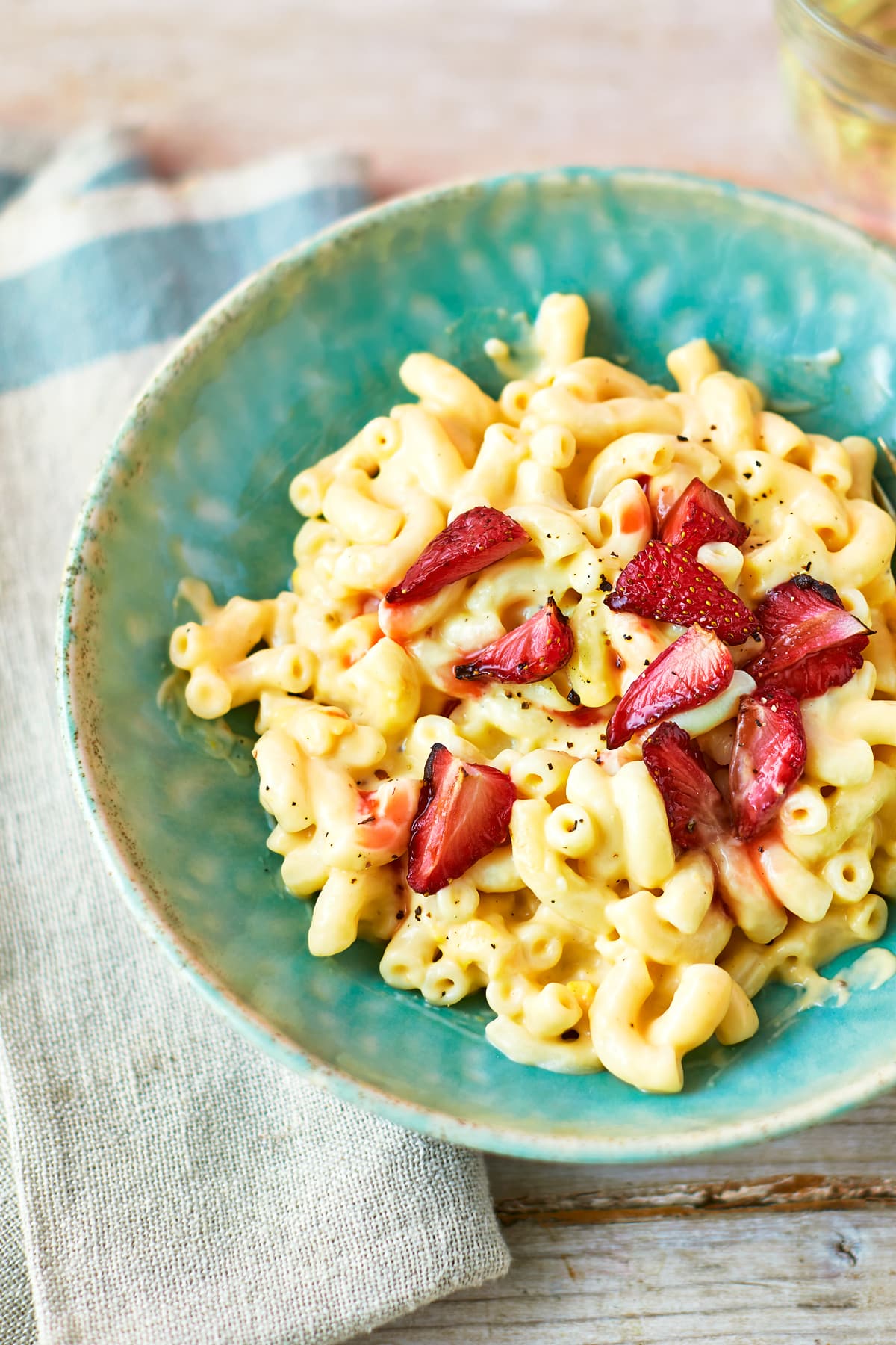 A turquoise bowl of strawberry pasta: macaroni in a cheese and butternut squash sauce, topped with roasted balsamic strawberries. The bowl rests on a tea towel on a wooden background.