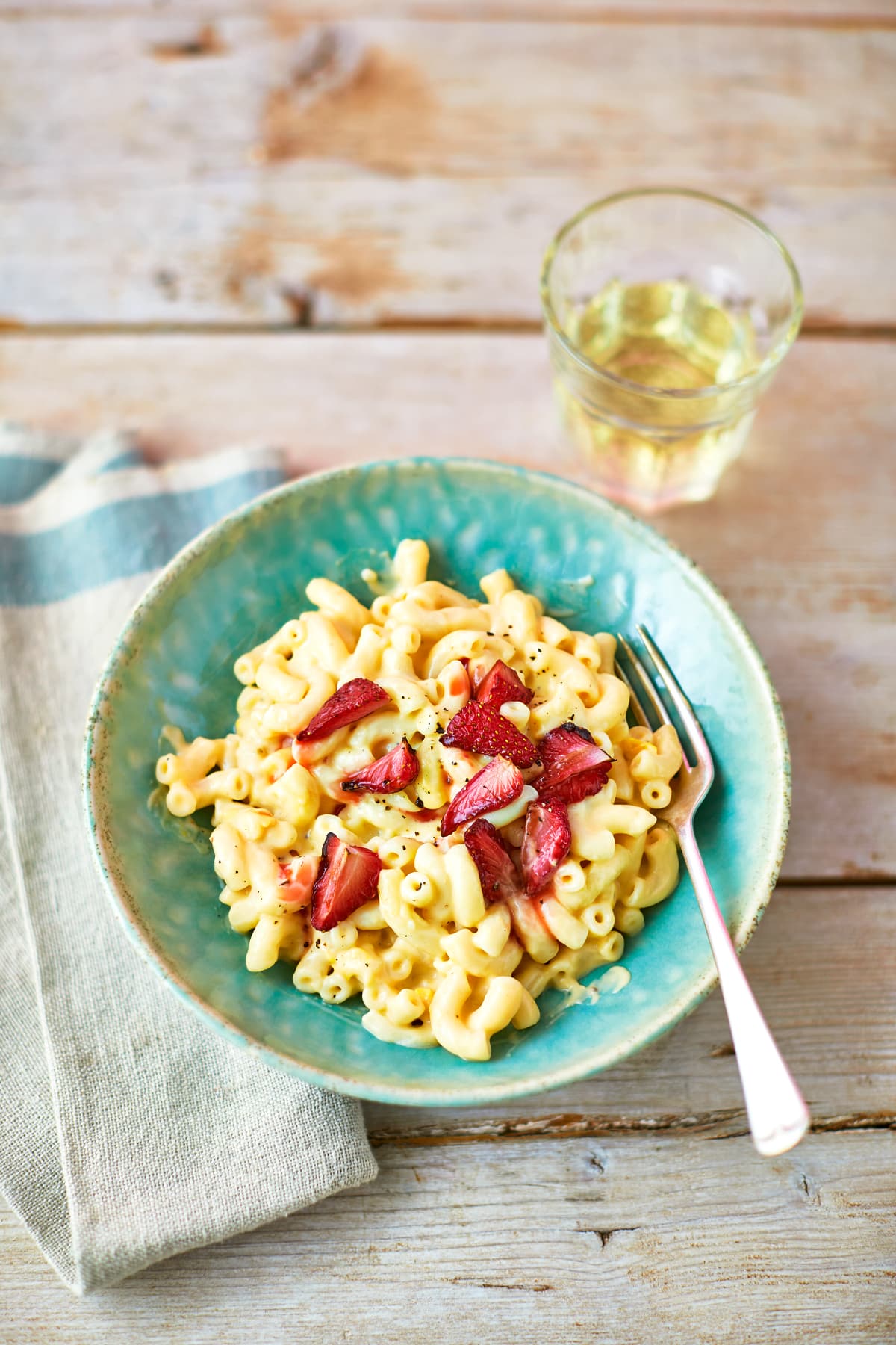 A turquoise bowl of strawberry pasta: macaroni in a cheese and butternut squash sauce, topped with roasted balsamic strawberries. A fork rests in the bowl by a glass of juice. The bowl sits on a tea towel on a wooden background.