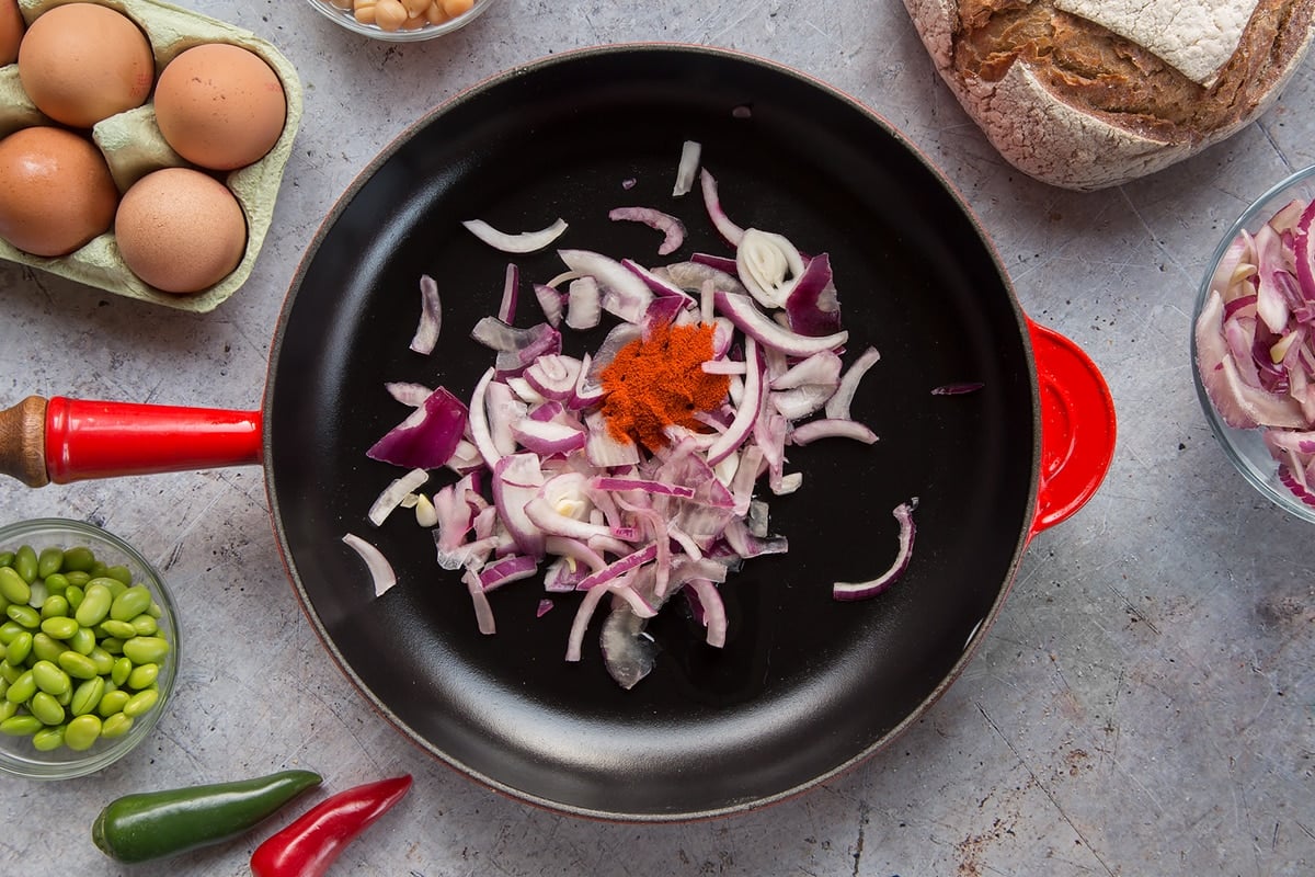 A pan with a little oil, sliced red onion and paprika. The pan is surrounded by ingredients to make breakfast beans.