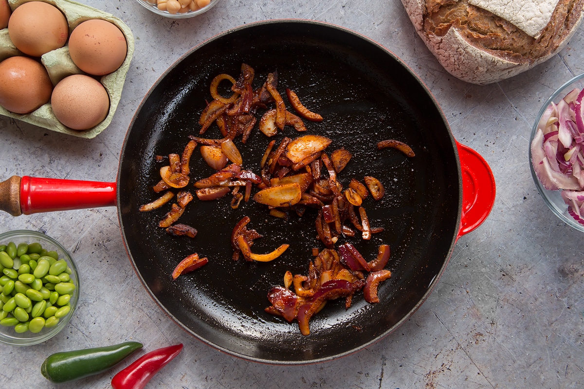 A pan with fried red onion and paprika. The pan is surrounded by ingredients to make breakfast beans.