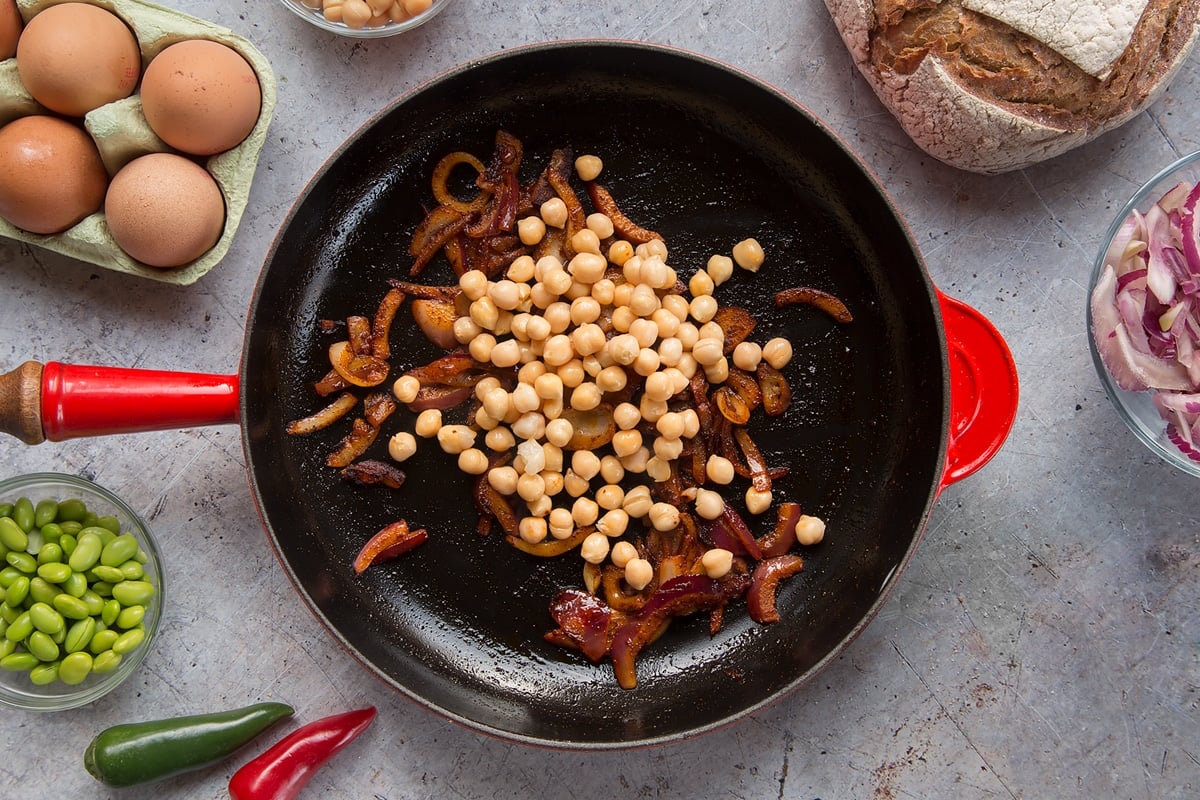 A pan with fried red onion and paprika. Chickpeas have been added. The pan is surrounded by ingredients to make breakfast beans.