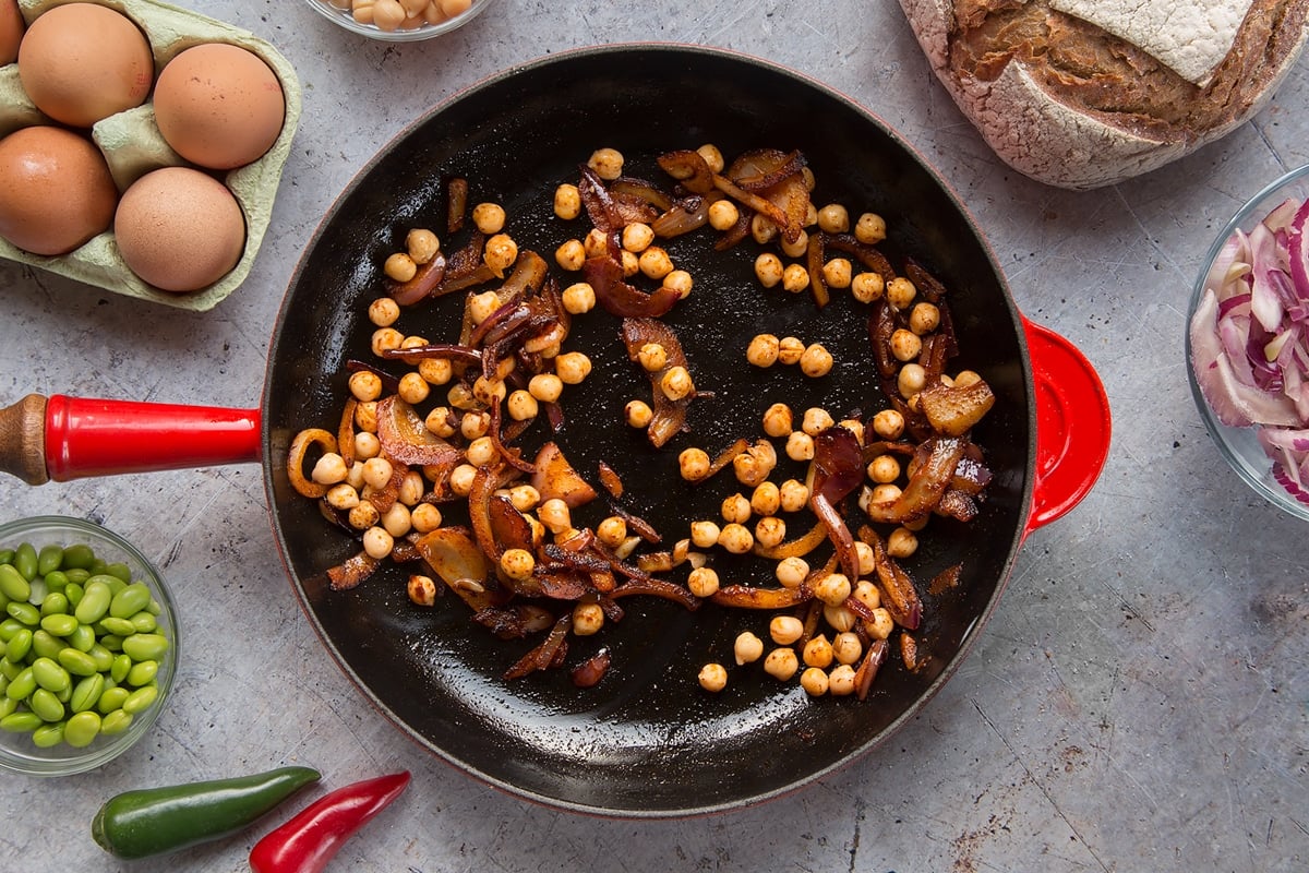 A pan with fried red onion, chickpeas and paprika. The pan is surrounded by ingredients to make breakfast beans.