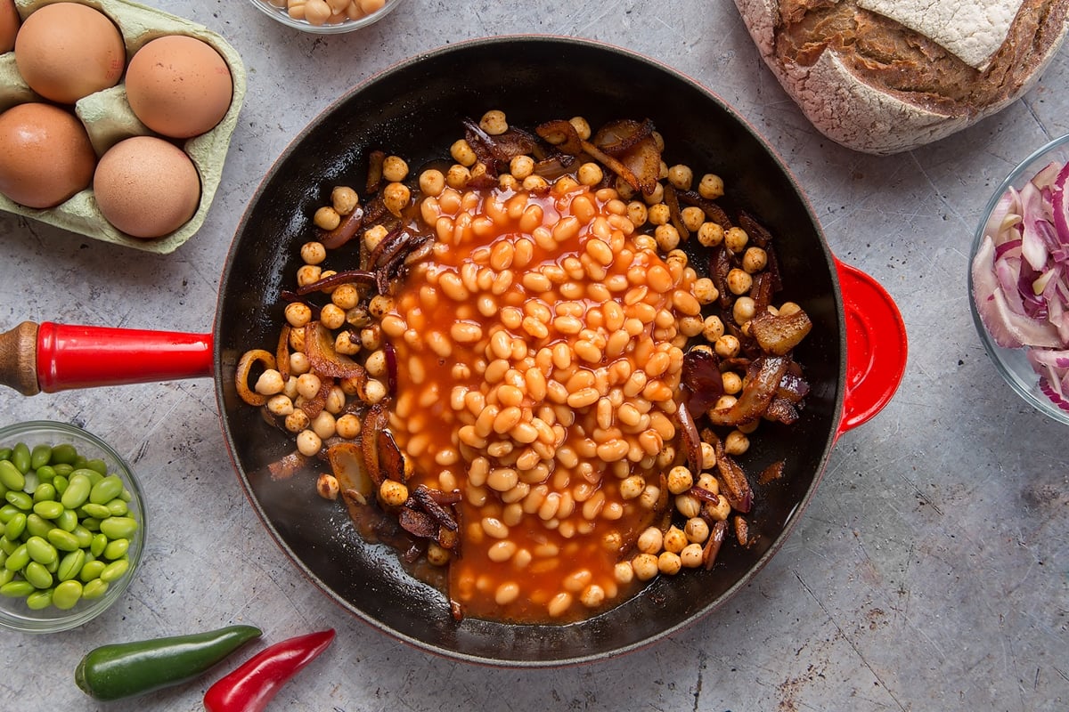 A pan with fried red onion, chickpeas and paprika. Baked beans have been added. The pan is surrounded by ingredients to make breakfast beans.