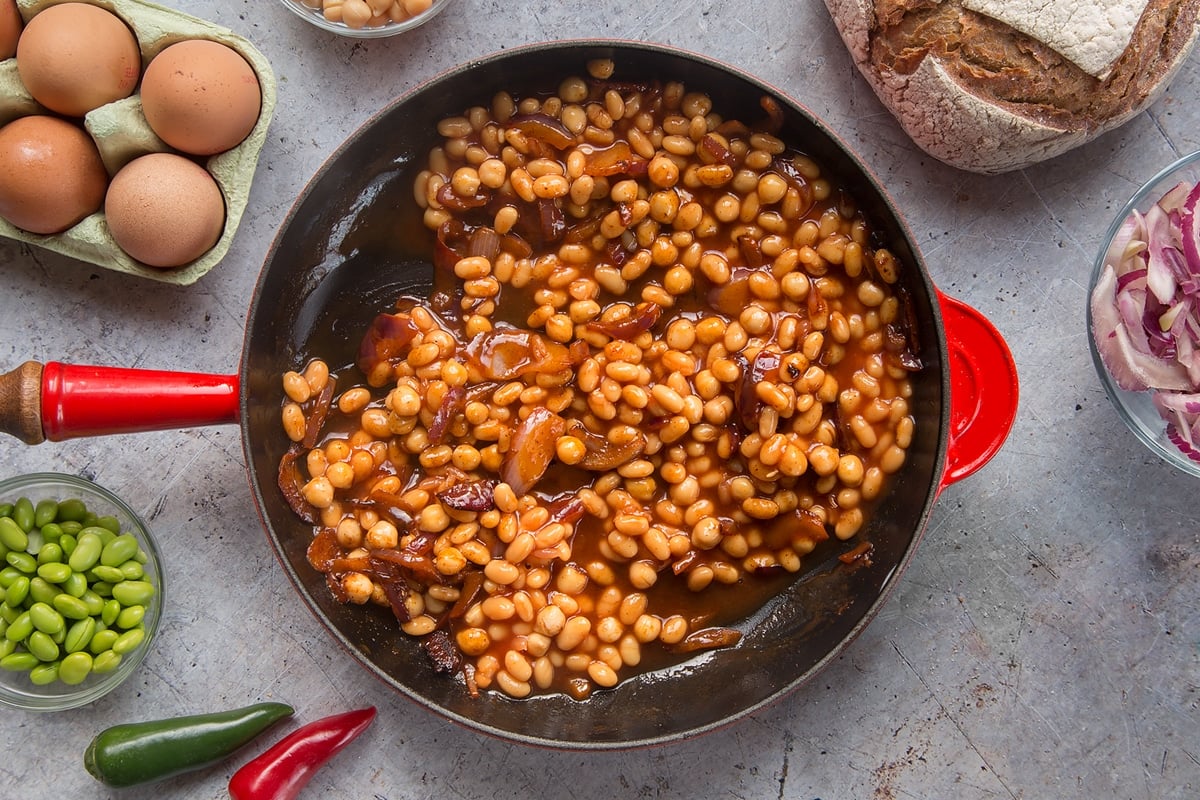 A pan with fried red onion, chickpeas, paprika and baked beans. The pan is surrounded by ingredients to make breakfast beans.