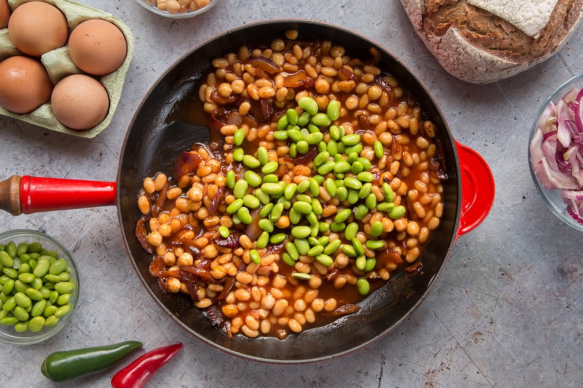 A pan with fried red onion, chickpeas, paprika and baked beans. Edamame have been added. The pan is surrounded by ingredients to make breakfast beans.