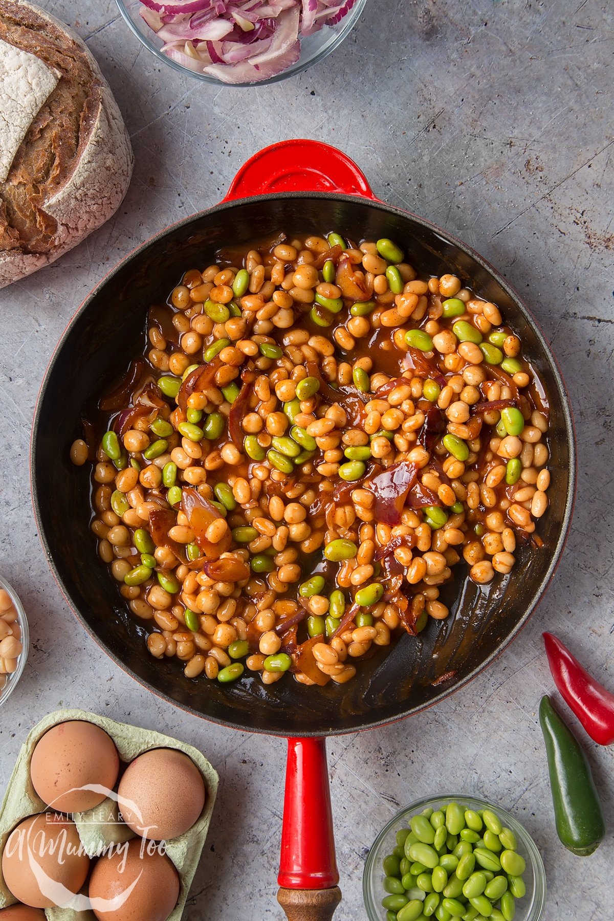 A pan with fried red onion, chickpeas, paprika, baked beans and edamame cooked together. The pan is surrounded by ingredients to make breakfast beans.