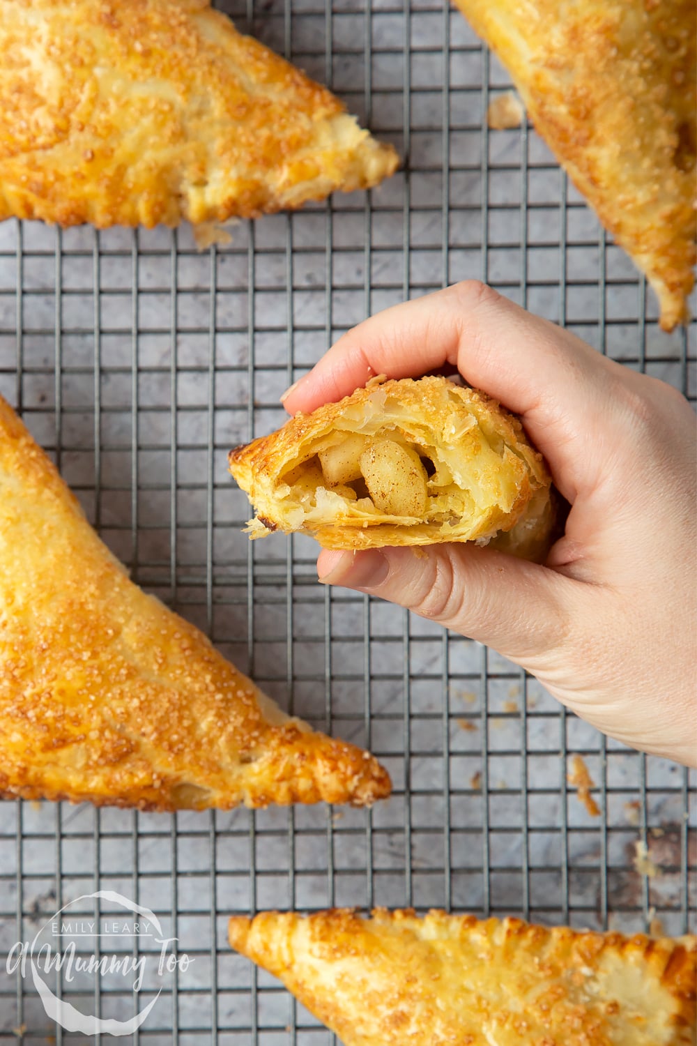 Overhead shot of a hand holding up an Apple cinnamon turnover on a baking rack with a mummy too logo in the lower-left corner.