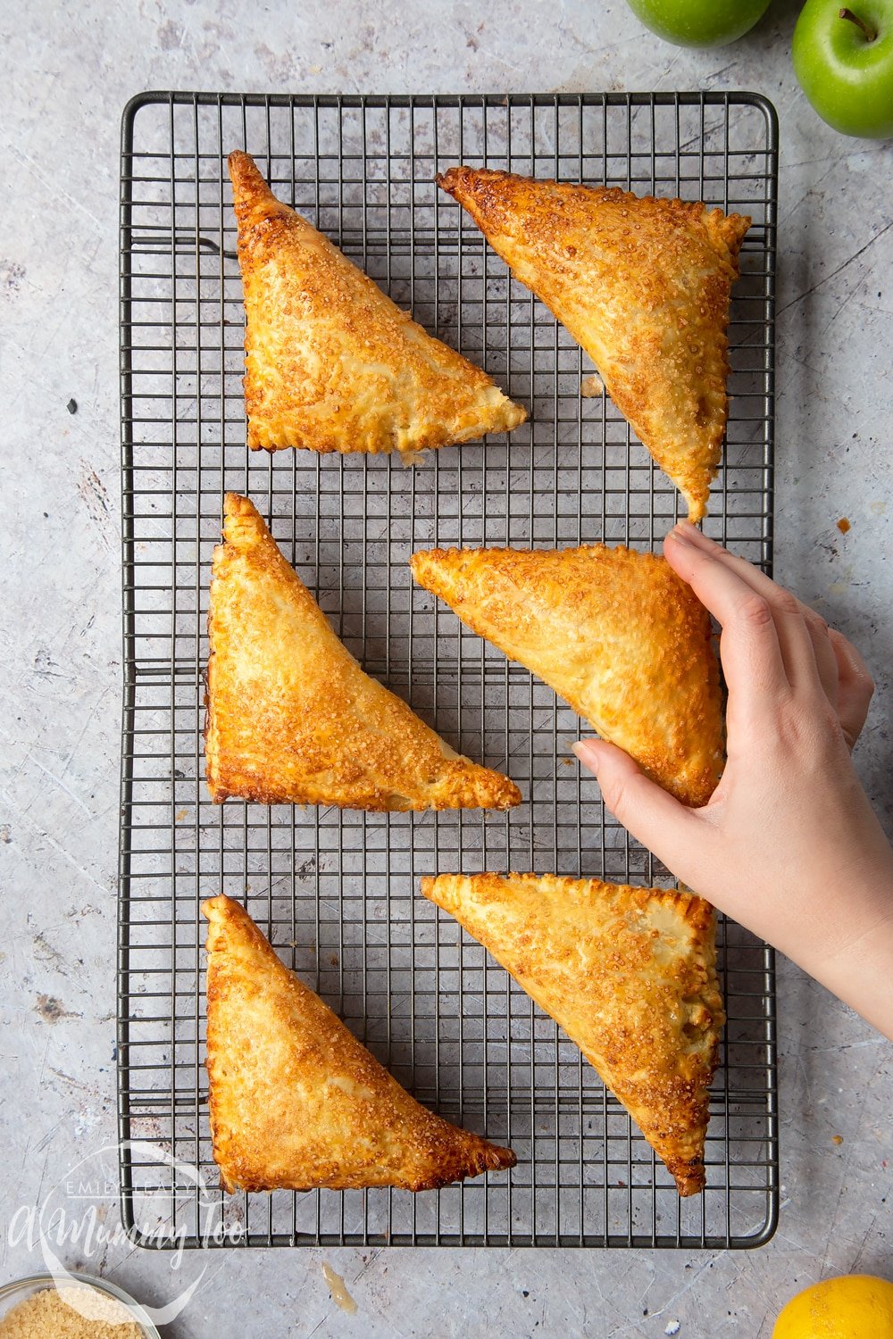 Overhead shot of a hand touching an Apple cinnamon turnover on a baking rack with other apple turnovers