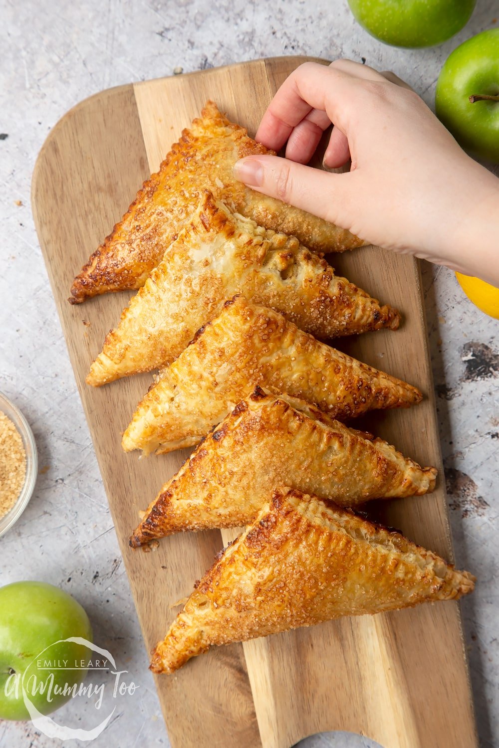 Five cinnamon apple turnovers lined up on a wooden chopping board with a hand holding one.