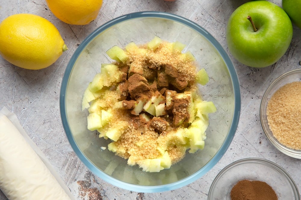 Overhead shot of chopped apples, cinnamon, and sugar in a clear bowl surrounded by sugar apples abd lemon