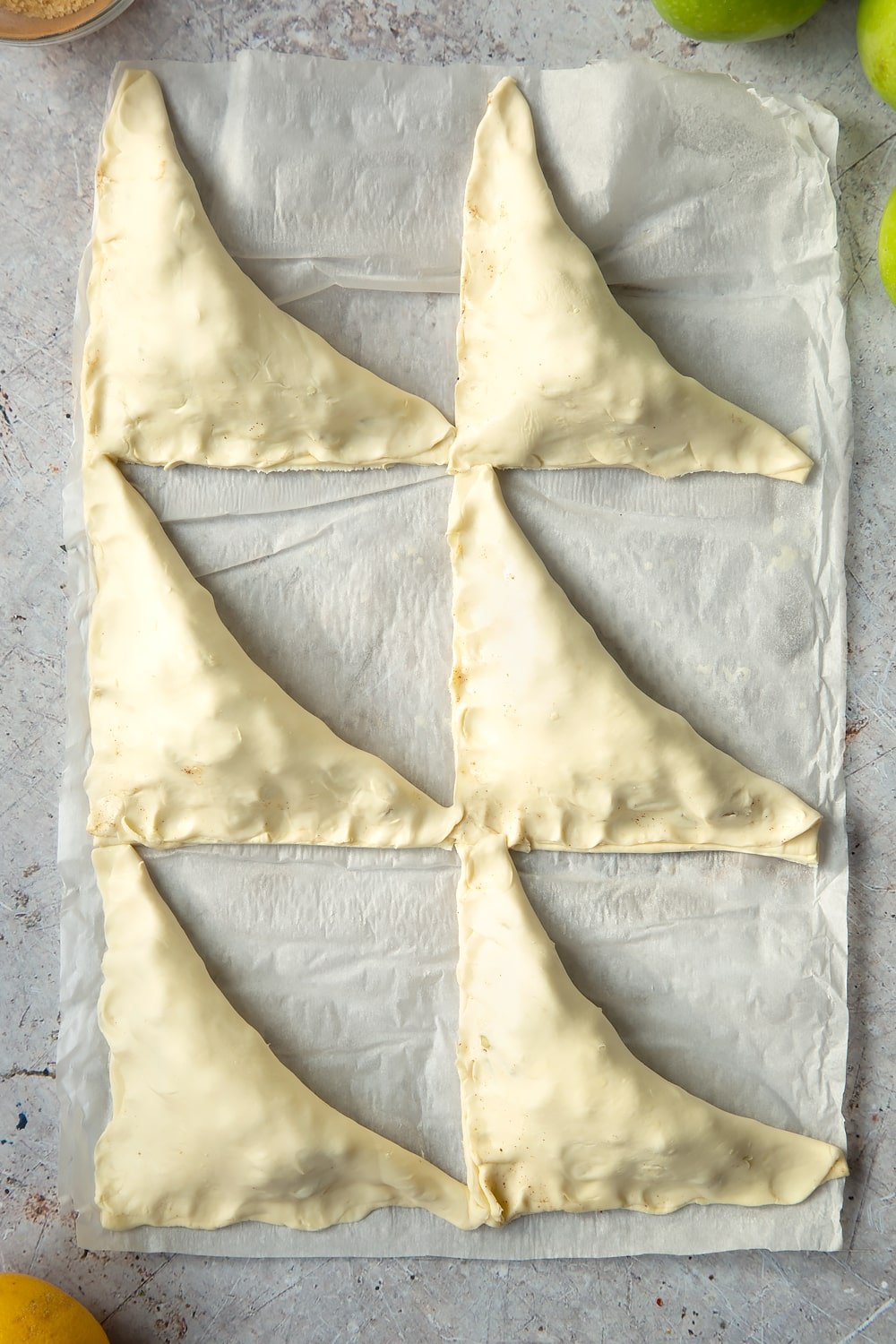 Overhead shot of 6 raw apple turnovers wrapped in pastry on a sheet of baking paper