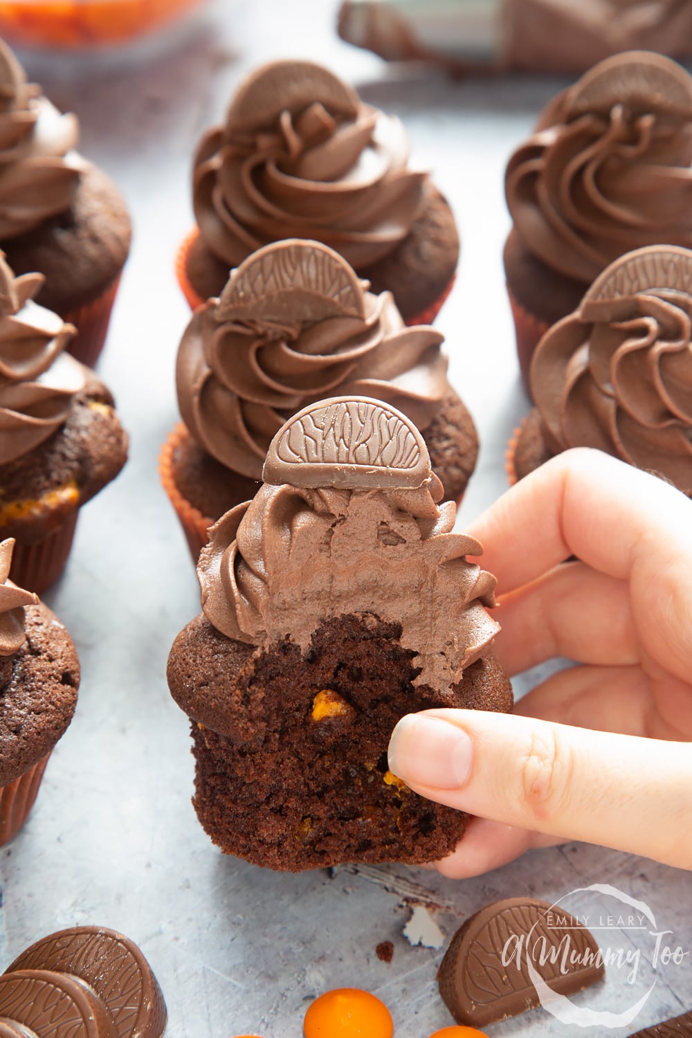 Front angle shot of a hand holding a chocolate orange muffin with a mummy too logo in the lower-right corner